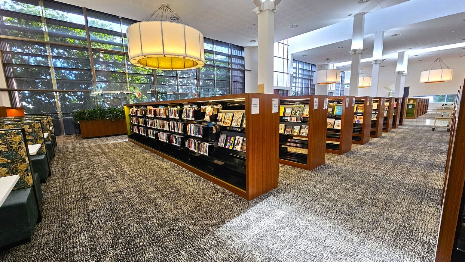 Library interior with rows of wooden bookshelves filled with books and magazines on the right. On the left, there are patterned upholstered seating booths with small tables. Large windows with a grid pattern allow natural light to filter in, casting shadows on the carpeted floor. Overhead, large circular pendant lights hang from the ceiling, which is white with recessed lighting. A planter with green foliage is positioned near the windows.