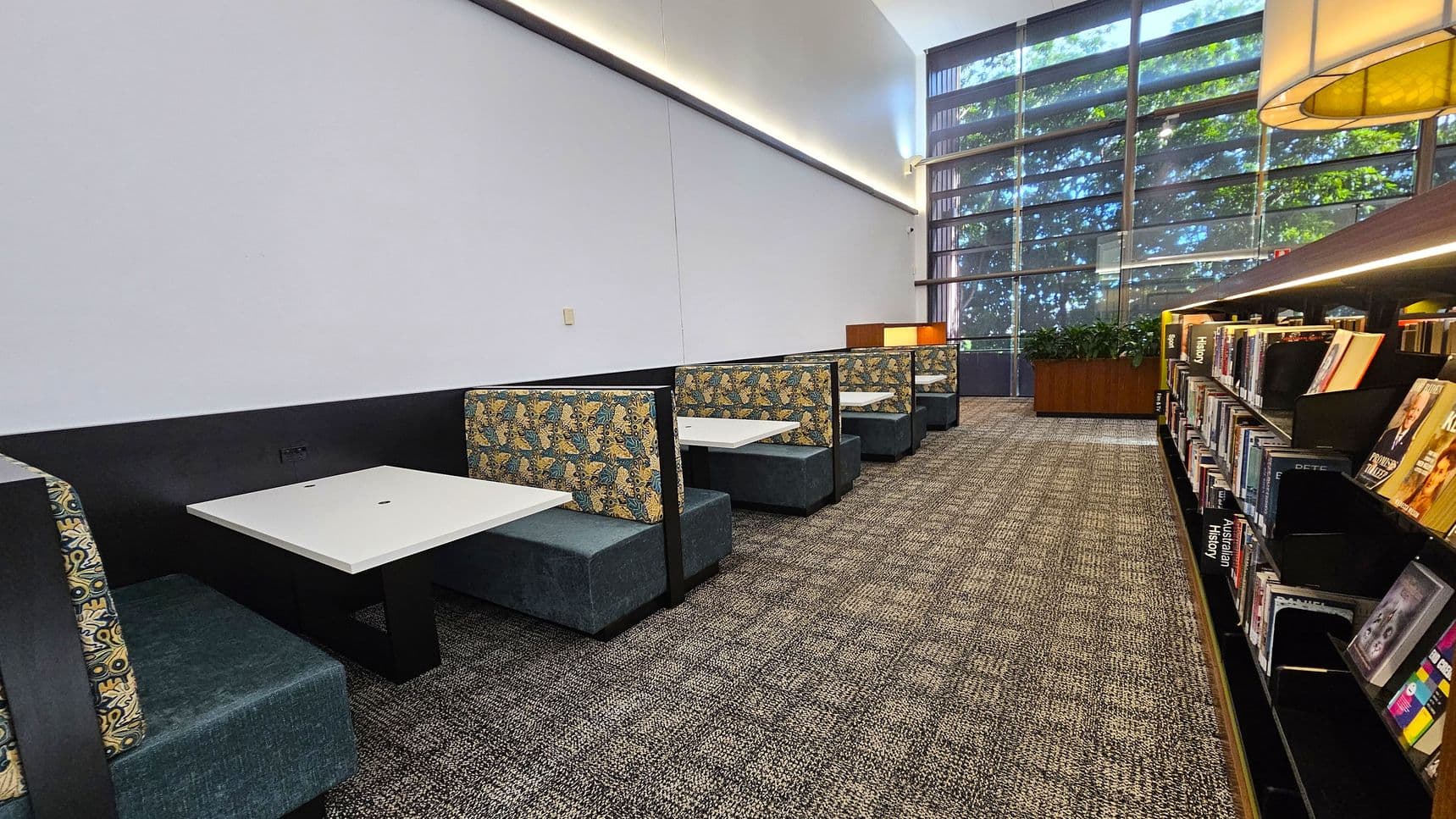 A library interior with a row of booth seating on the left, featuring patterned upholstery and white tables. The right side has bookshelves filled with books. The floor is carpeted with a textured pattern. Large windows in the background allow natural light, with greenery visible outside. A planter with green plants is positioned near the windows. The ceiling is high with a strip of lighting along the wall.