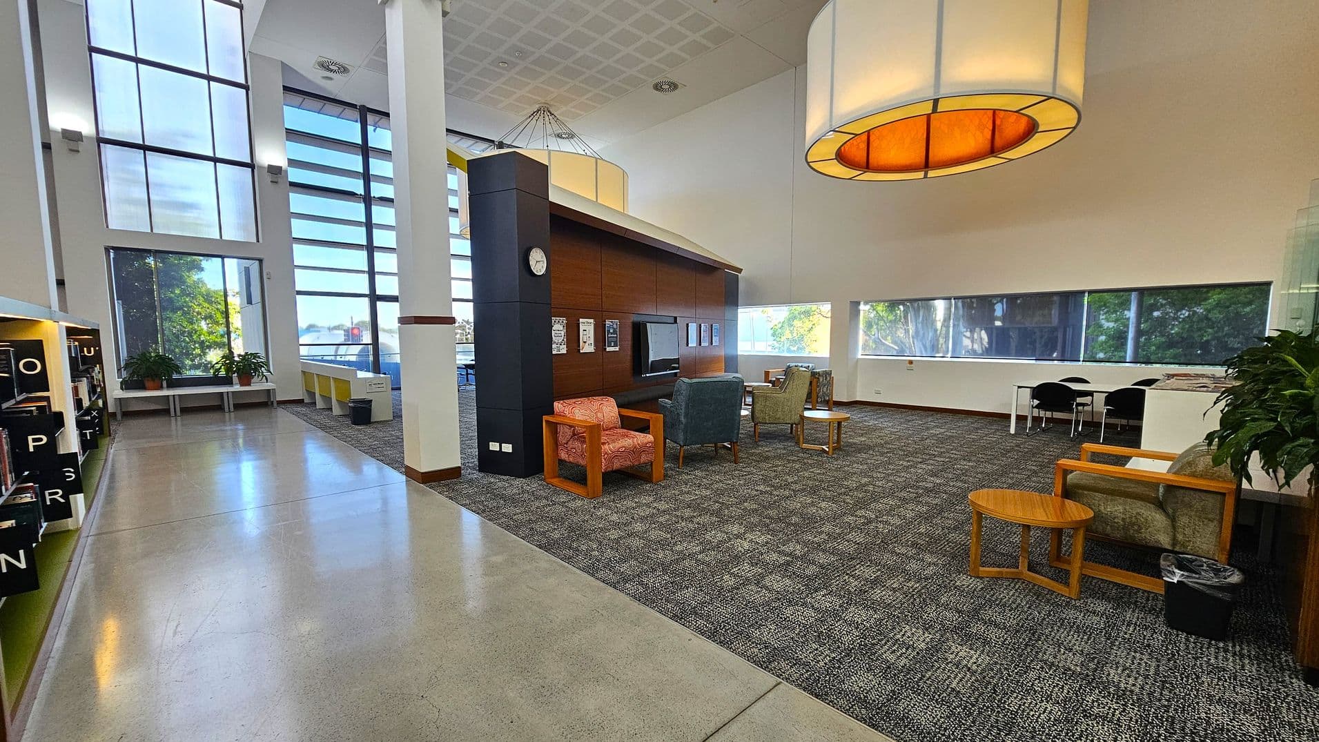 A spacious interior with a high ceiling and large windows on the left, allowing natural light to flood in. The left side features bookshelves with labelled sections and potted plants on a bench. In the centre, there is a seating area with various chairs and small tables on a carpeted floor. A wooden panelled wall with a clock and notices is present. On the right, there are more chairs, tables, and a long window with a view of trees. A large, circular ceiling light hangs above.