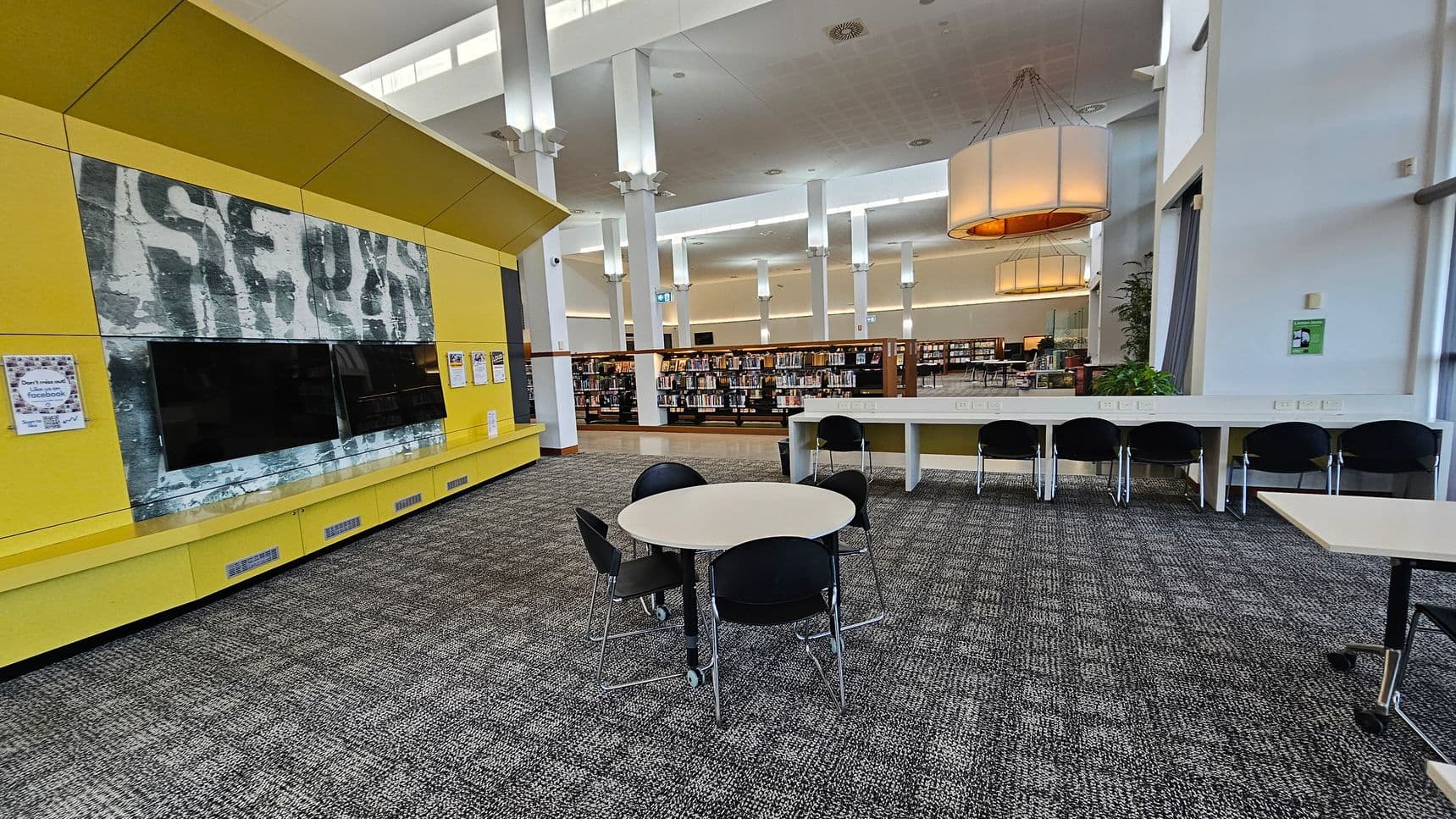 A library interior with a yellow feature wall on the left, displaying a large screen and some posters. In the centre, a round table with four black chairs is placed on a patterned carpet. On the right, a long white desk with several black chairs is positioned against the wall. In the background, bookshelves filled with books are visible, along with tall white columns and large hanging light fixtures. The ceiling is high with recessed lighting.