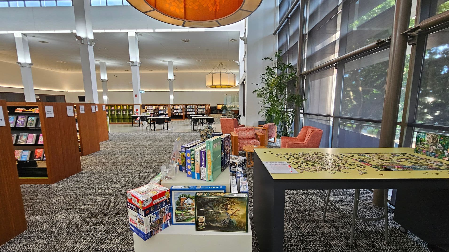 A spacious library interior with a high ceiling and large windows on the right, allowing natural light to filter in. In the foreground, a table with a partially completed jigsaw puzzle and several puzzle boxes. To the right, two orange armchairs and a small table are near a potted plant. On the left, rows of wooden bookshelves filled with books. In the background, more seating and bookshelves are visible, with a large circular light fixture hanging from the ceiling.