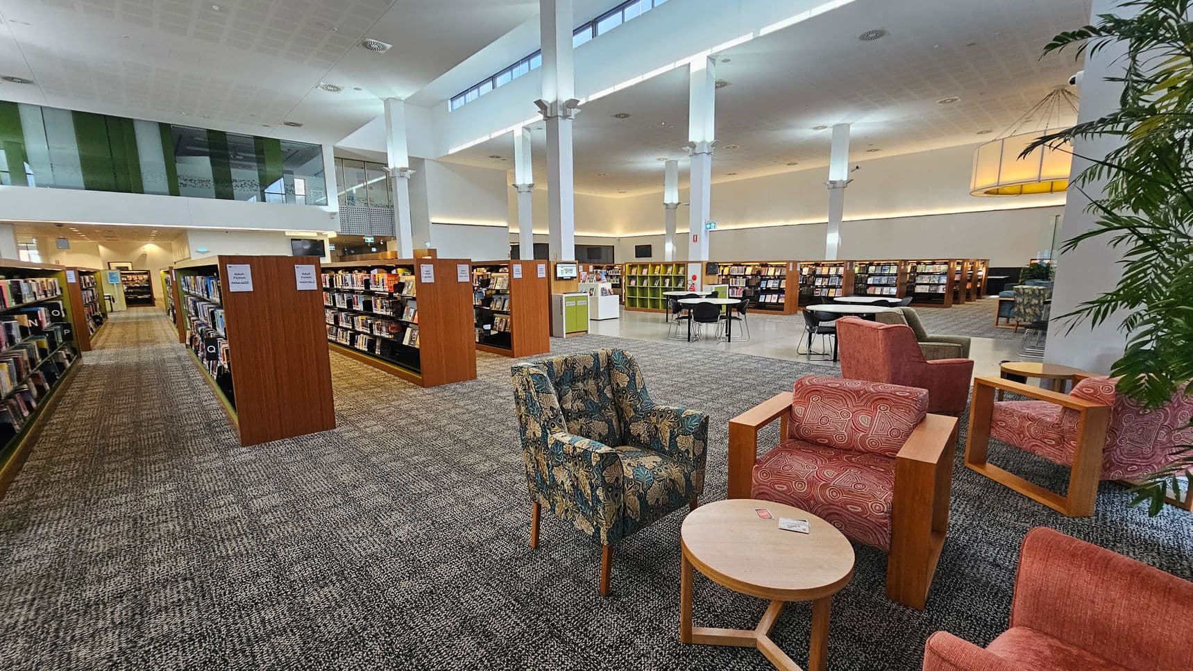 A spacious library interior with bookshelves arranged in rows on the left and centre. In the foreground, there are patterned and solid-coloured armchairs around a small round table. The floor is carpeted with a grey and green pattern. The ceiling is high with white pillars and overhead lighting. In the background, there are more bookshelves and a seating area with tables and chairs. A large plant is visible on the right.