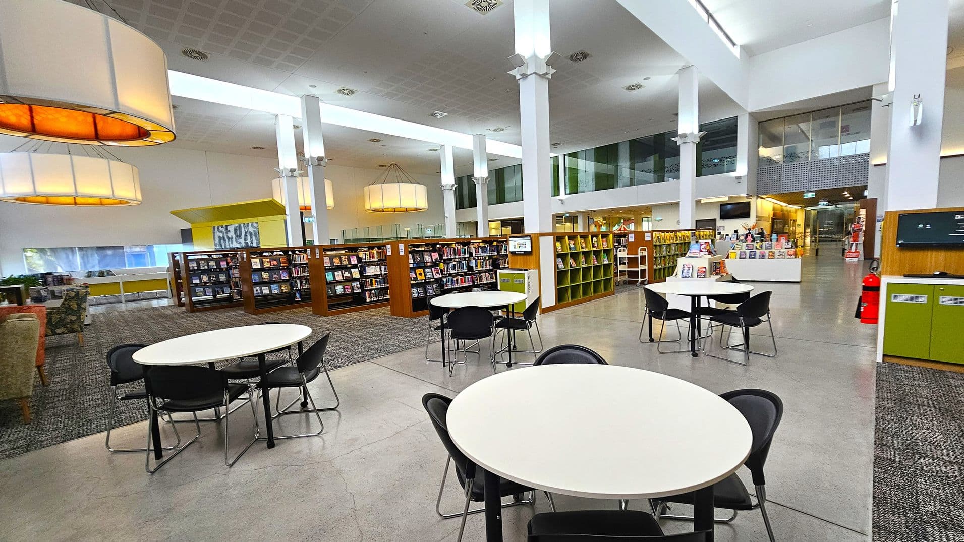 A spacious library interior with a high ceiling and large, circular hanging lights. In the foreground, there are round tables with black chairs. The middle section features bookshelves filled with books, and a section with a yellow sign. The background has a reception area with a display of books and a fire extinguisher nearby. The floor is a mix of carpet and polished concrete, and the walls are white with large windows allowing natural light.