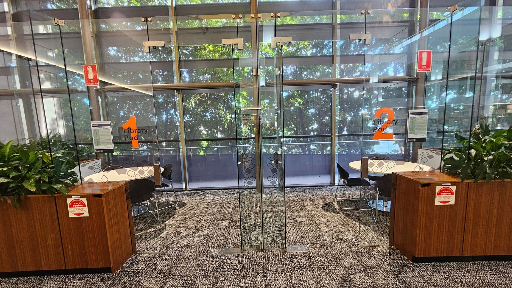 Glass doors in the centre lead to two library pods, numbered 1 and 2 in orange. Each pod has a round table and chairs. On the left and right, there are wooden cabinets with green plants on top. Fire extinguisher signs are visible above the doors. The background features large windows with a view of trees outside. The floor is carpeted in a grey pattern.