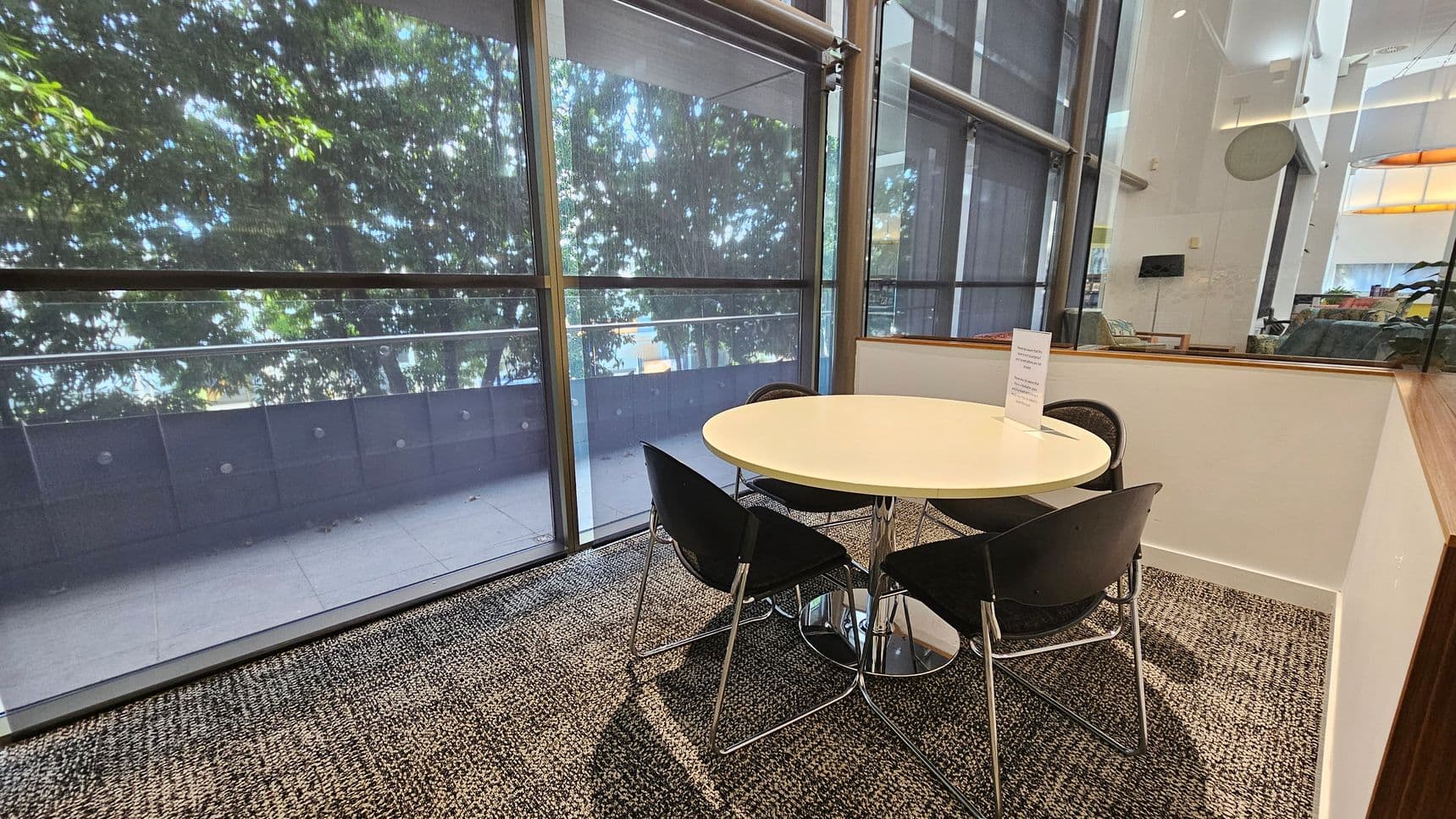 A modern indoor seating area with a round white table surrounded by four black chairs with metal legs. The floor is covered in a patterned carpet. Large glass windows on the left reveal a view of trees outside, partially obscured by blinds. On the right, a glass partition separates this area from a larger room, which contains more seating and a wall-mounted screen. The ceiling is high with visible lighting fixtures.