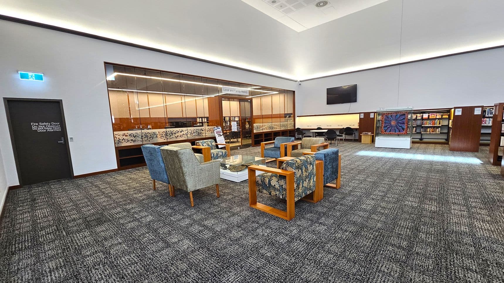 A spacious library interior with a grey carpeted floor. On the left, there's a dark door marked as a fire safety exit. In the centre, a seating area features cushioned chairs with wooden frames around a glass table. Behind, a glass-walled room labelled \"Local and Family History\" is visible. On the right, bookshelves line the wall, and a display case with a colourful artwork is present. The ceiling is high with recessed lighting.