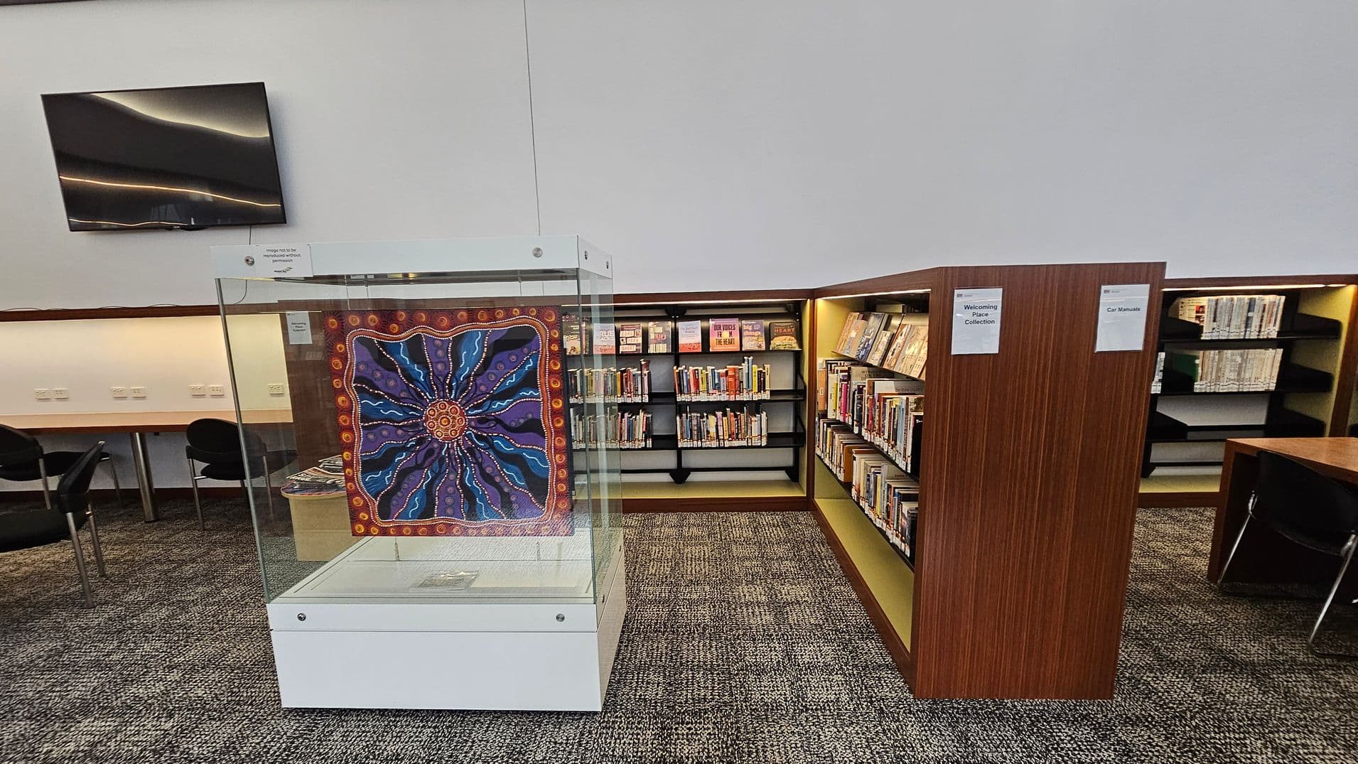 A library interior with a glass display case in the centre featuring a colourful artwork with a circular pattern. To the right, there are wooden bookshelves filled with books, labelled \"Welcoming Place Collection\" and \"Car Manuals\". On the left, a row of chairs and desks is visible against the wall. Above, a large, dark rectangular panel is mounted on the wall. The floor is carpeted with a patterned design.