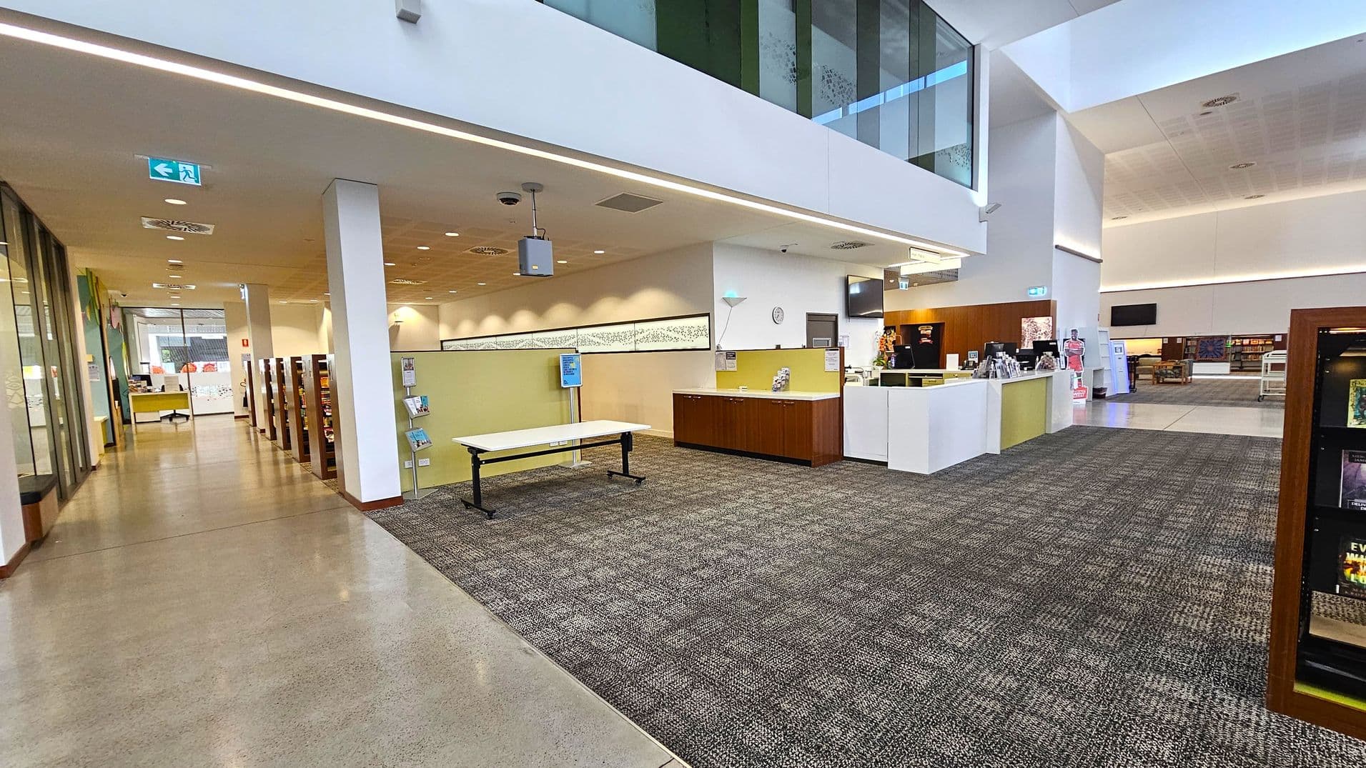 A spacious library interior with a high ceiling. On the left, there is a corridor with bookshelves and a desk near the entrance. In the centre, a reception area with a counter and a clock on the wall. A table and a display stand are nearby. The floor is a mix of carpet and polished concrete. On the right, more bookshelves and a seating area are visible. The upper level has glass panels, and the lighting is bright and even throughout.