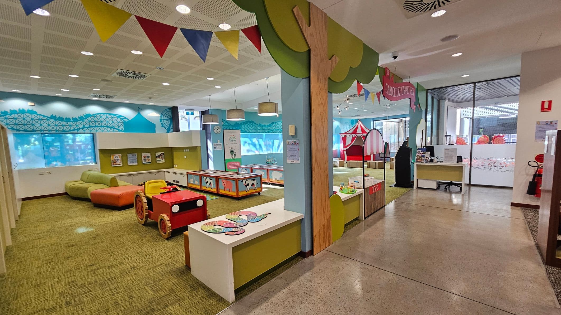 A colourful children's play area with a green and orange sofa on the left, a red toy car, and a play table with cow designs. The walls have a blue mural with a roller coaster and tent. On the right, there's a circus tent structure and a desk with a computer. The ceiling has colourful bunting and round lights. The floor is a mix of carpet and polished concrete. Large windows in the background let in natural light.