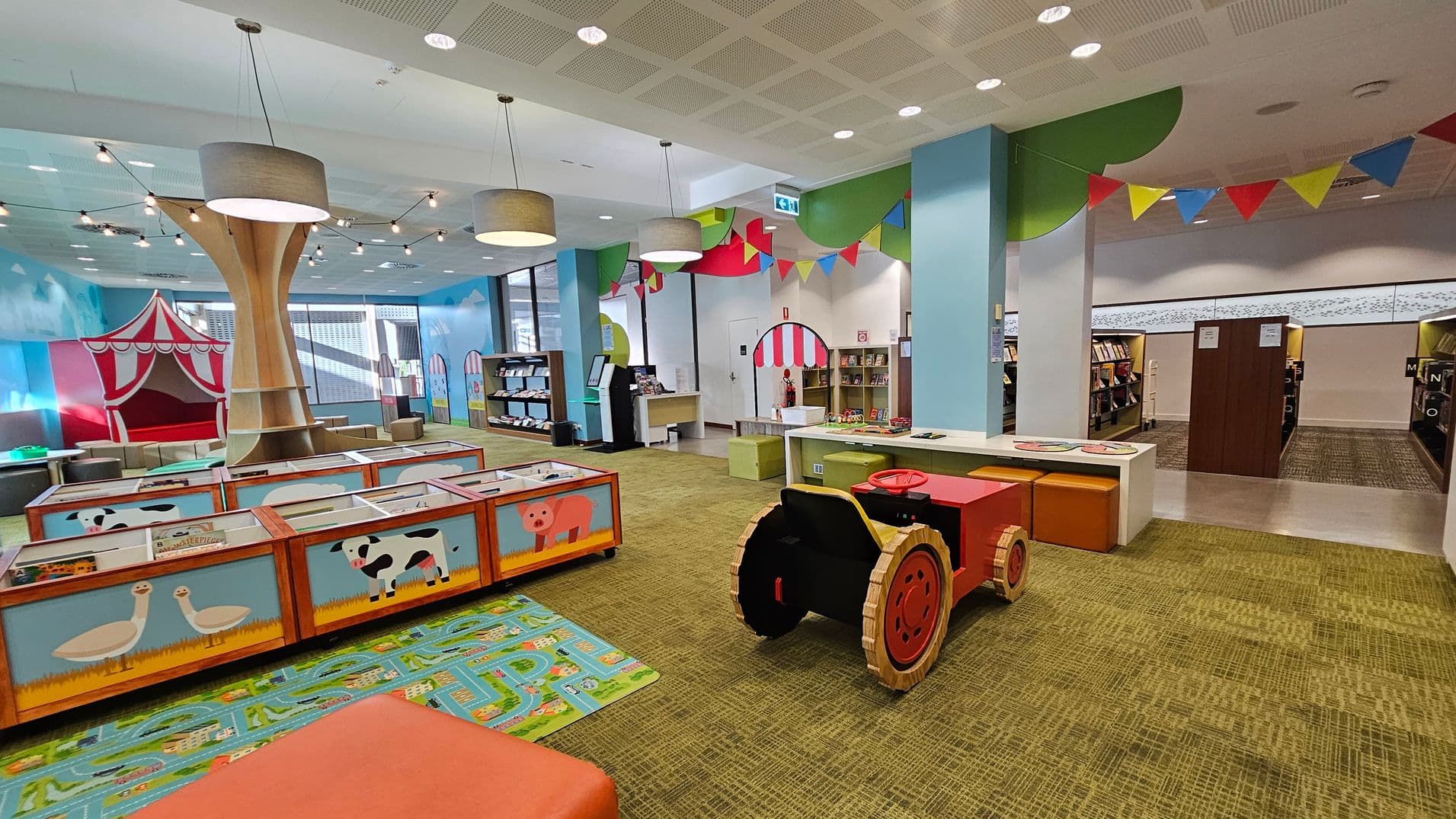 A colourful children's library area with a playful design. On the left, there are low bookshelves with animal illustrations and a large wooden tree structure. In the centre, a red and black toy tractor sits on a patterned carpet. The right side features a small table with stools and bookshelves in the background. Brightly coloured bunting hangs from the ceiling, and the walls are painted with whimsical scenes, including a red and white circus tent. The floor is carpeted in a green pattern.