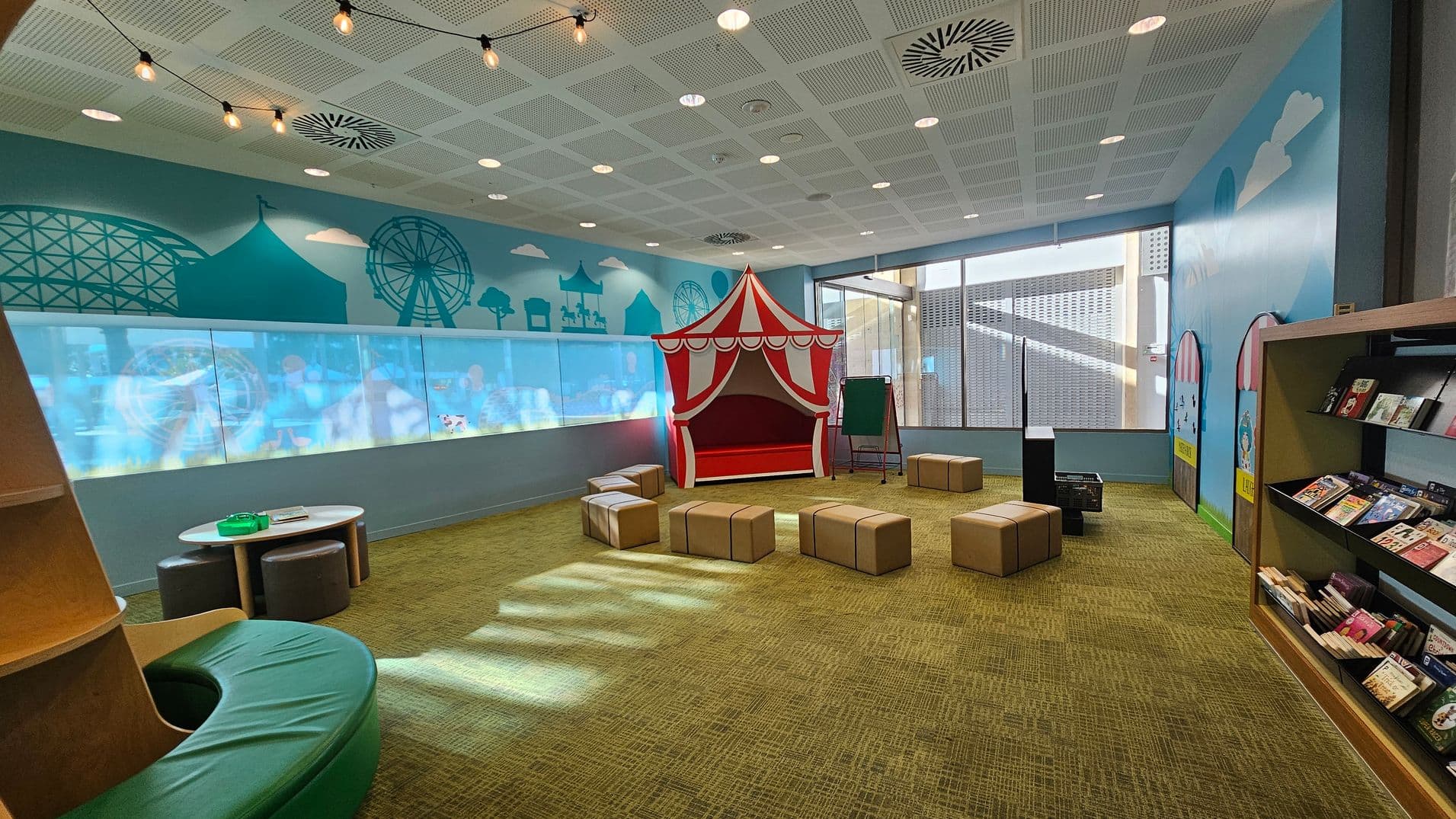 A children's playroom with a circus theme. On the left, there's a circular green cushioned seating area. In the centre, a red and white striped tent is surrounded by cube-shaped stools. The right side features a bookshelf filled with books. The walls are decorated with illustrations of a Ferris wheel and roller coaster. The ceiling has a grid pattern with recessed lights and hanging bulbs. The floor is carpeted in a greenish-brown tone. Large windows allow natural light to enter.