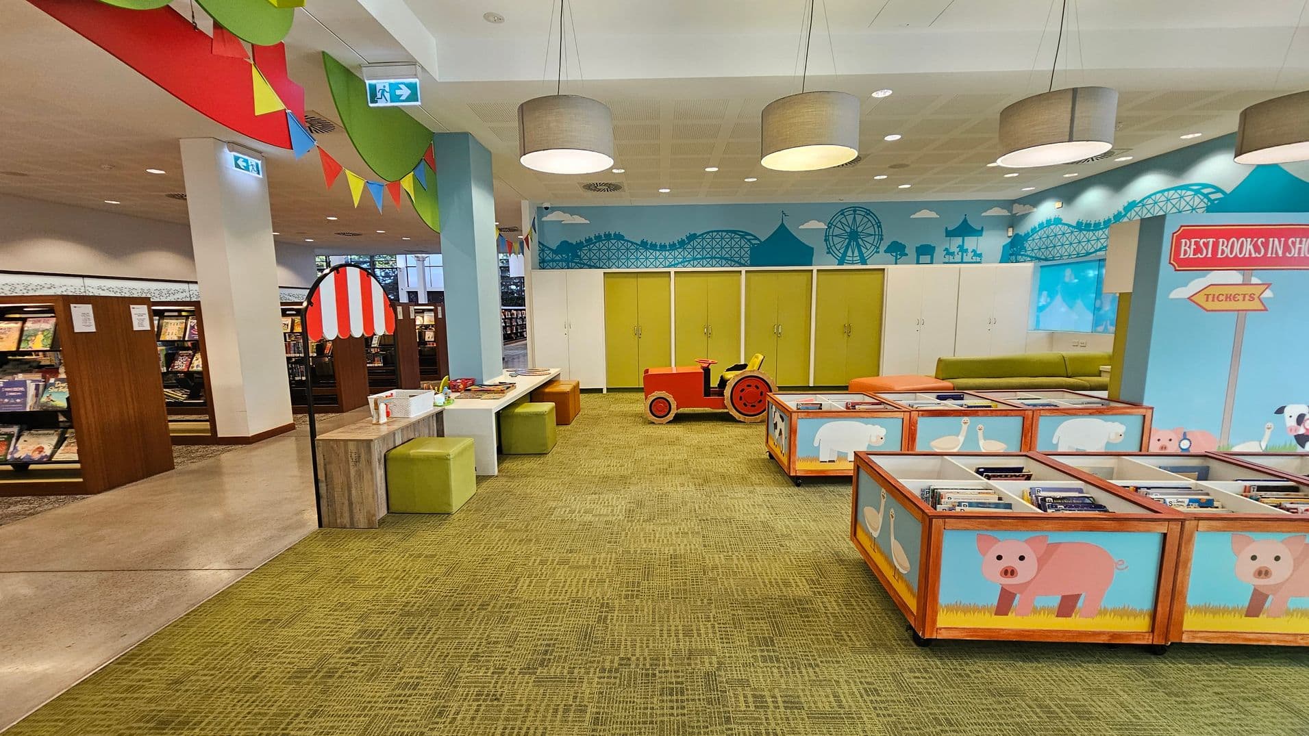 A children's library area with a playful theme. On the right, there are colourful book bins decorated with animal illustrations like pigs and ducks. In the centre, a red toy tractor sits on a green carpet. The back wall features a mural of a fairground with a Ferris wheel and roller coaster. On the left, bookshelves are filled with books. The ceiling has large circular lights, and colourful bunting hangs across the space.