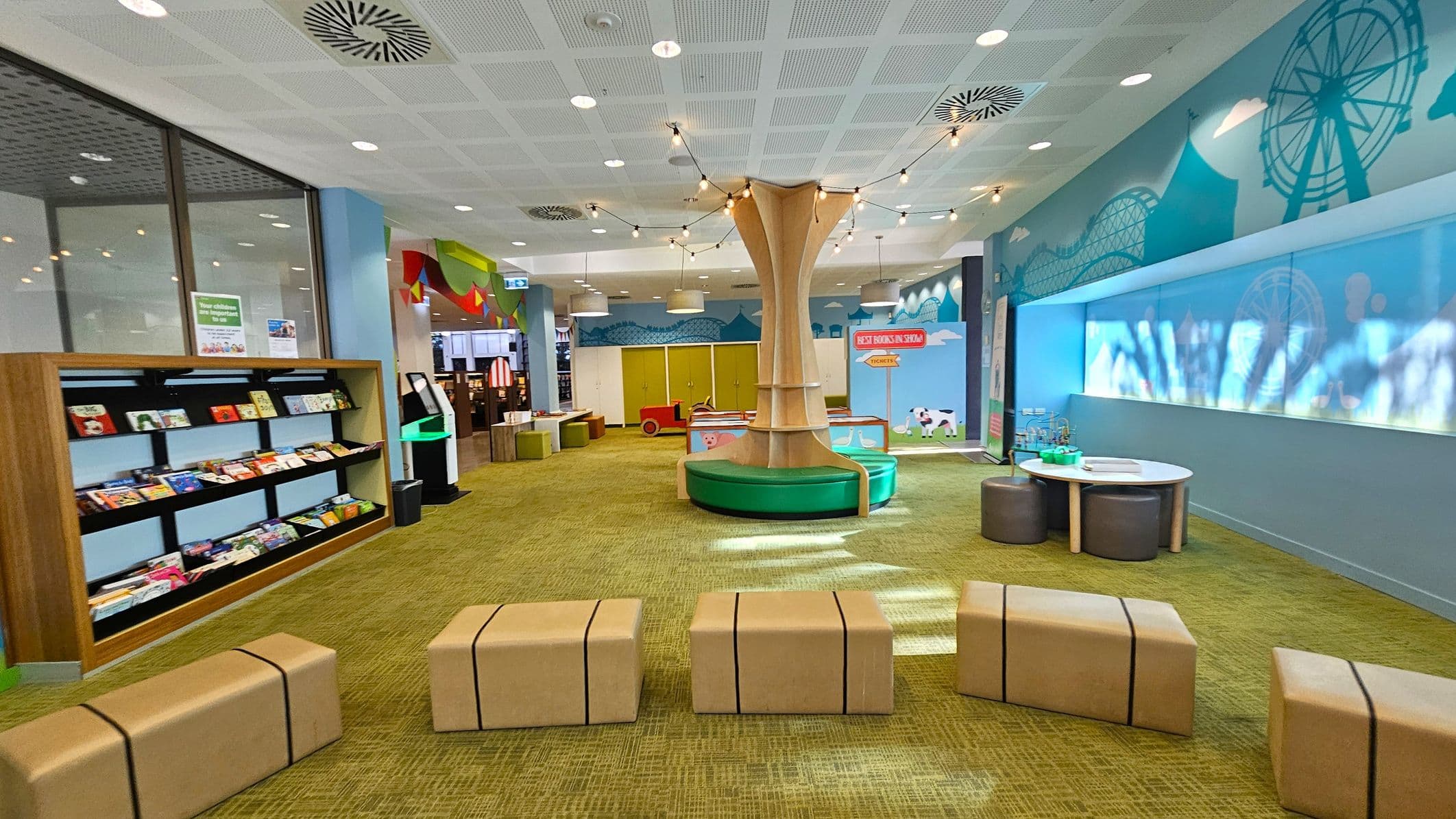 A children's play area with a green carpeted floor. On the left, there is a bookshelf filled with colourful books. In the centre, a large wooden structure resembling a tree with seating around its base is adorned with string lights. To the right, a wall features a mural of a Ferris wheel and roller coaster, with a small round table and stools beneath it. The ceiling is white with circular lights and air vents.