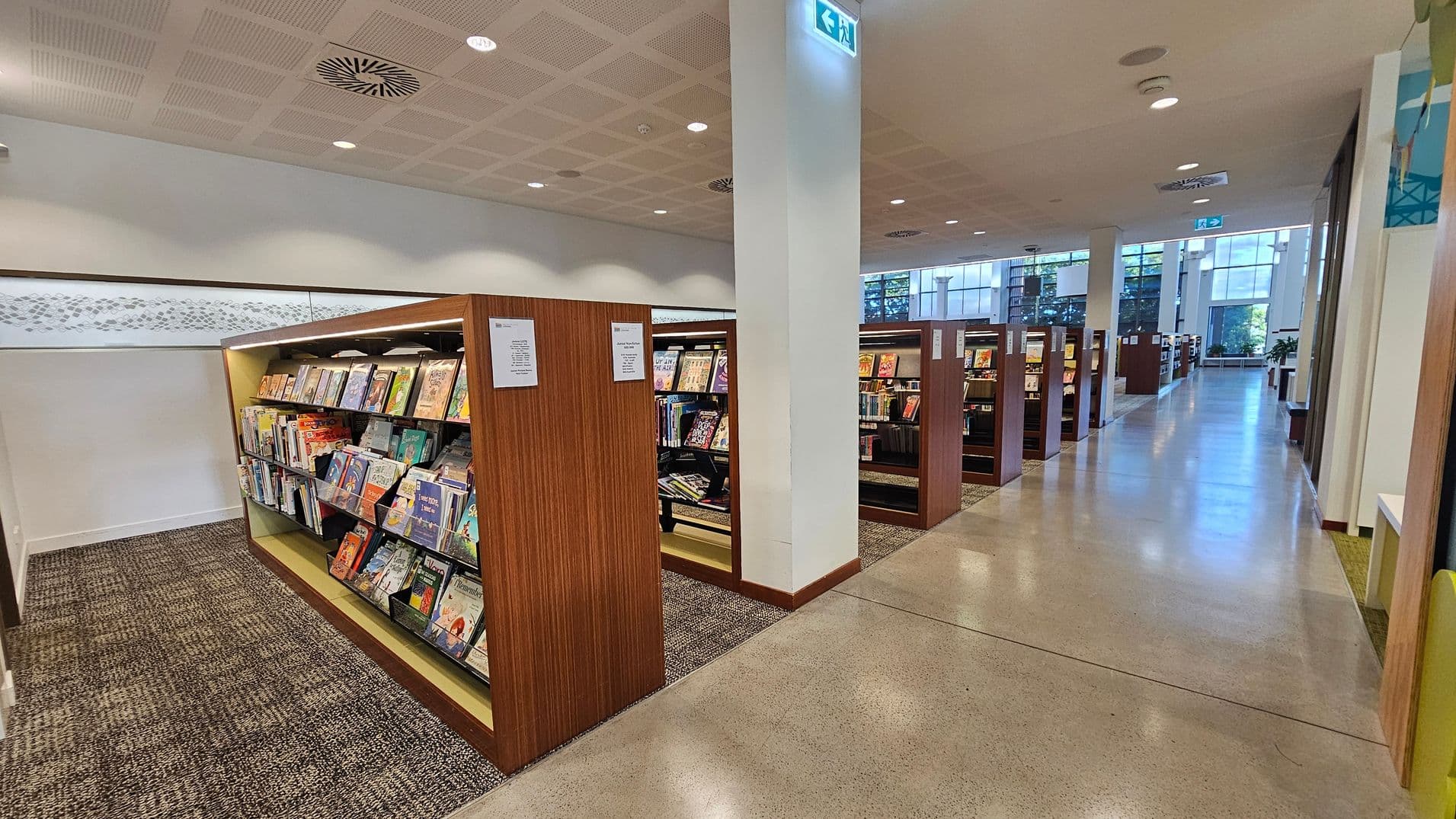 Bookshelves filled with colourful books are on the left, with a carpeted floor beneath them. To the right, a polished concrete floor leads down a long aisle lined with more bookshelves. Large windows in the background allow natural light to fill the space. The ceiling is fitted with recessed lighting and ventilation panels. An exit sign is visible above a white column.