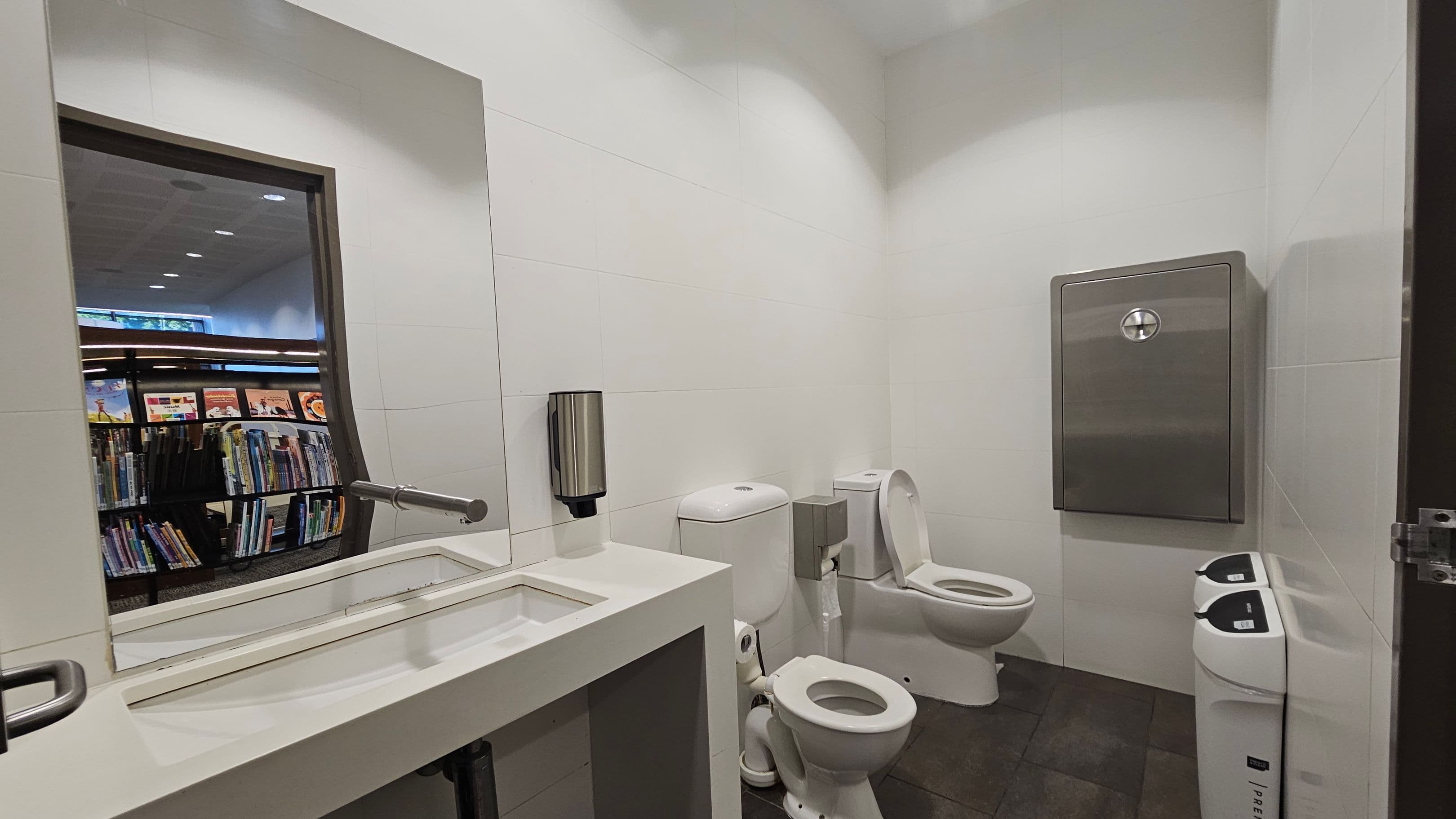 A public restroom with a white sink and a mirror on the left wall. The mirror reflects a library section with books on shelves. Below the mirror is a soap dispenser. On the right, there are two toilets, one adult-sized and one child-sized, with a metal toilet paper holder between them. A stainless steel waste bin is mounted on the wall above two white bins on the floor. The floor is dark grey tile, and the walls are covered in white tiles.