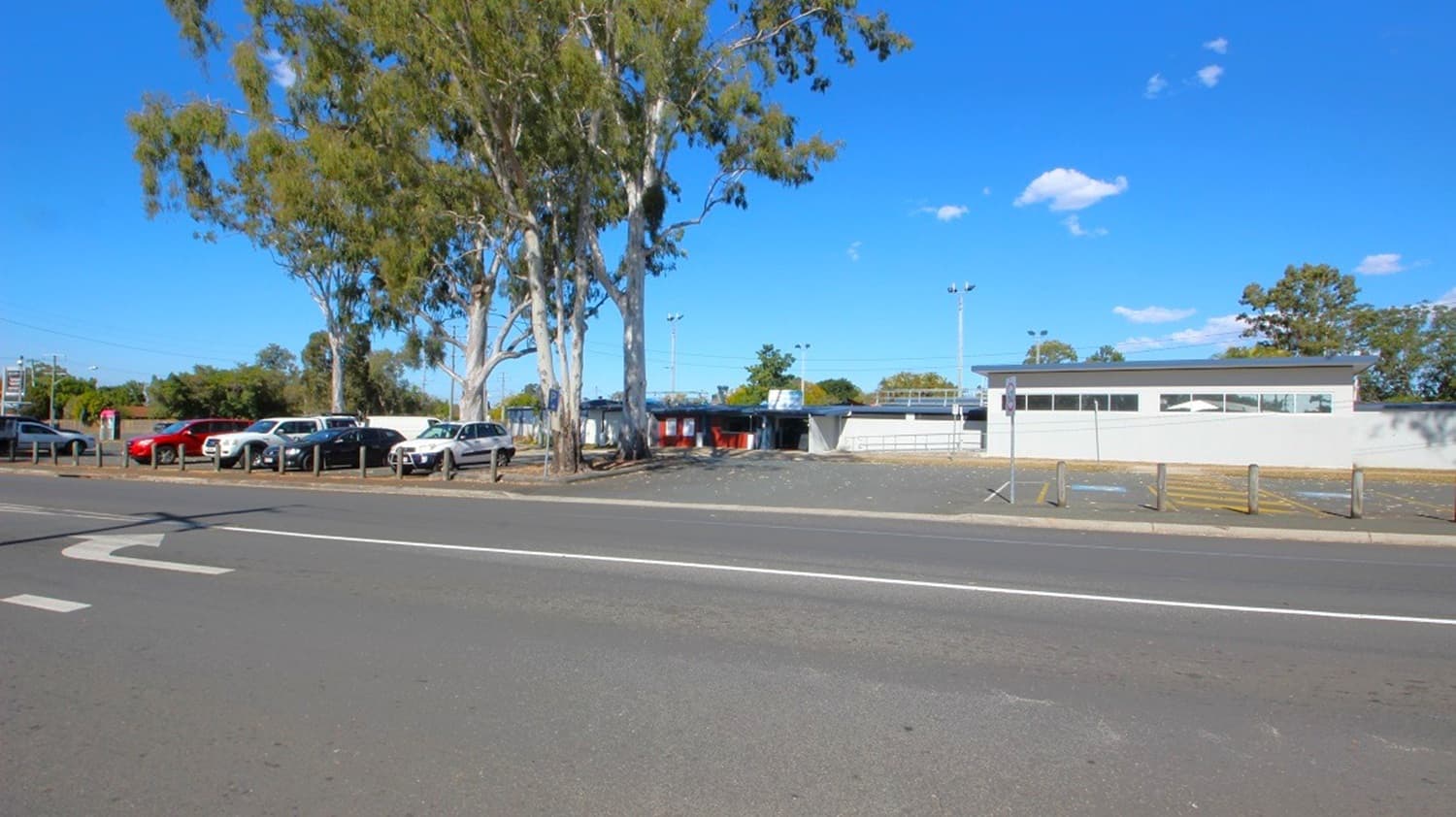 A row of parked cars is on the left, next to a line of tall trees. In the centre, there is a small building with a flat roof and several windows. The foreground features a wide road with a white arrow painted on it. On the right, there is a grassy area with a few scattered trees. The background includes more trees and a clear blue sky.