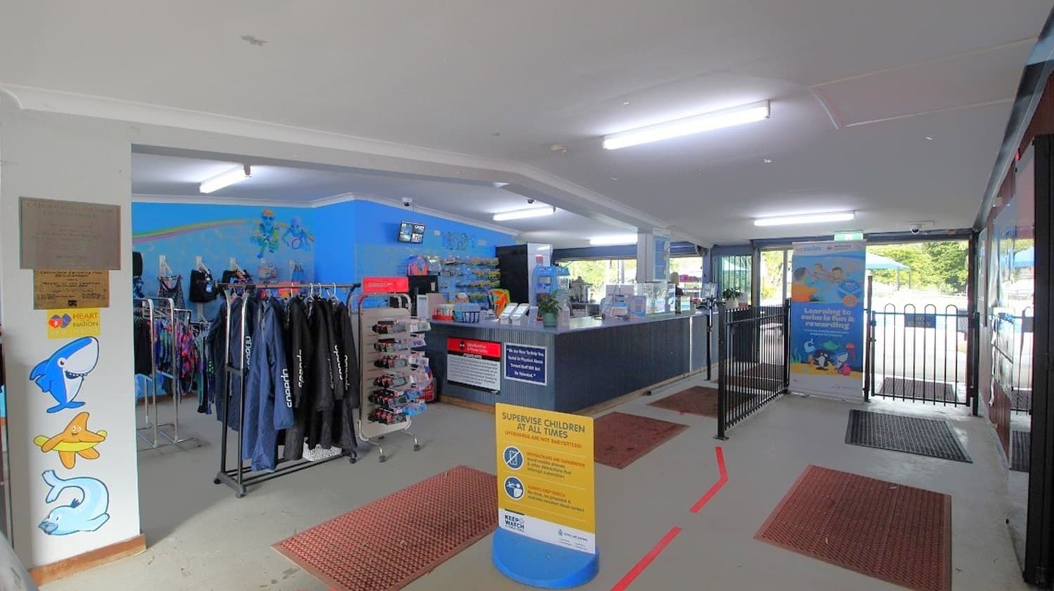 Inside a shop with a blue-themed interior. On the left, there are racks of wetsuits and swim gear. In the centre, a counter displays various items with a sign about supervising children. On the right, a gated entrance leads outside, with a large poster nearby. The floor is grey with red mats, and the ceiling has fluorescent lights.