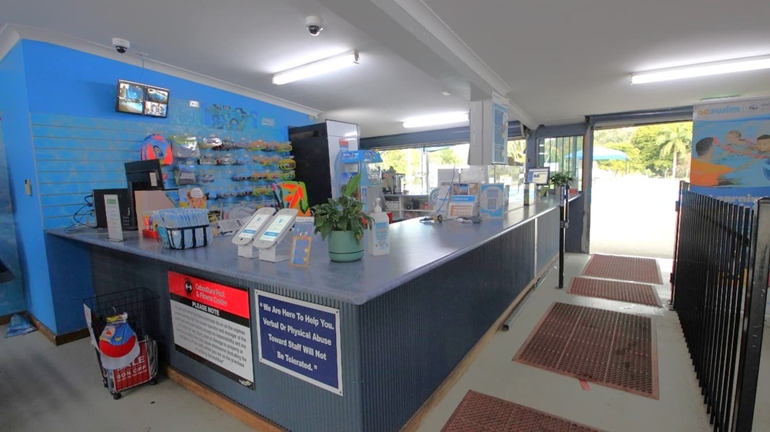 Reception area with a long counter on the right, featuring brochures, a plant, and hand sanitiser. Behind the counter, a blue wall displays various items for sale. On the left, a small TV screen is mounted above. The floor has brown mats leading to a glass entrance on the right, showing a glimpse of greenery outside. The ceiling is white with fluorescent lights.