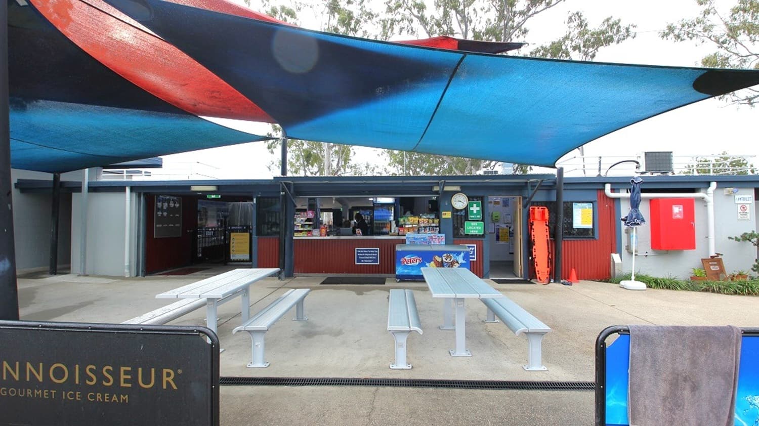 A small outdoor café with a counter in the centre, displaying snacks and drinks. Above, there are large blue and red shade sails. In front, several long, grey picnic tables with benches are arranged on a concrete floor. On the right, a red emergency box and a life preserver are mounted on the wall. Trees are visible in the background.