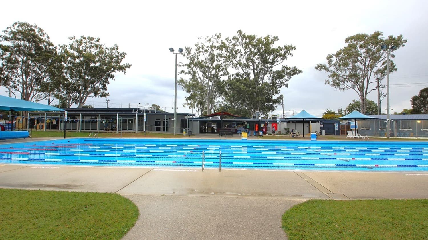 Outdoor swimming pool with blue and white lane dividers in the centre. Surrounding the pool is a concrete walkway and patches of green grass. In the background, there are several tall trees and a row of low buildings, possibly changing rooms or facilities. A few blue shade structures are visible on the left and right sides. The sky is overcast, and there are tall light poles around the area.