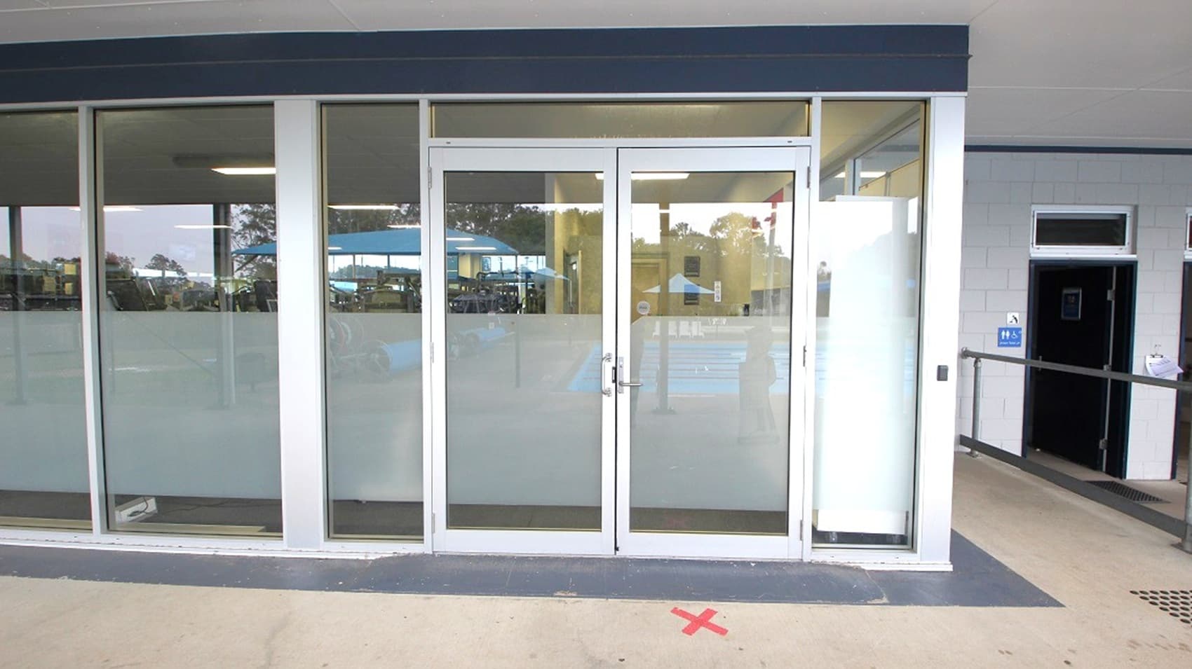 Glass entrance with double doors in the centre, framed by large windows on either side. The lower half of the glass is frosted. Inside, fluorescent lights and reflections of an interior space are visible. To the right, a corridor with a ramp and a sign indicating accessibility. The floor is light-coloured with a red "X" mark in front of the doors. The ceiling is plain and light-coloured.