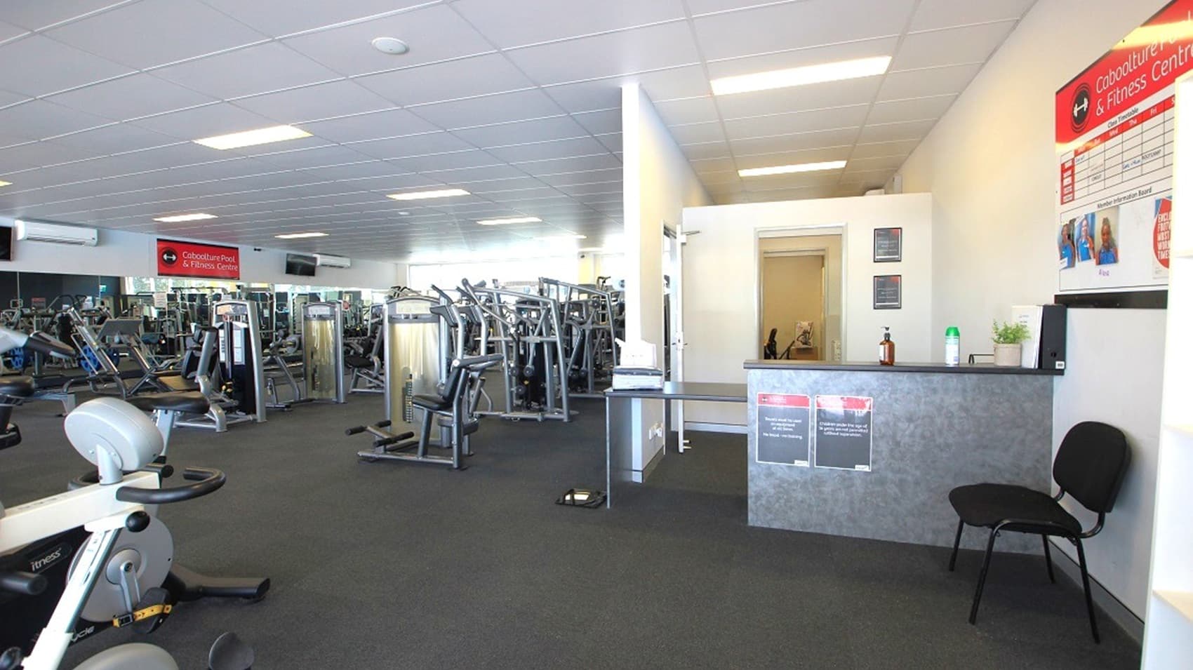 A gym interior with a reception area on the right, featuring a counter, a chair, and wall posters. In the middle, there's an open space leading to a room with more equipment. On the left, numerous exercise machines are arranged in rows. The floor is carpeted, and the ceiling has recessed lighting.