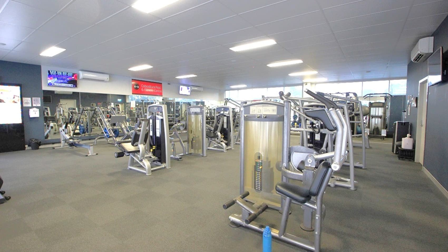 A spacious gym interior with various exercise machines arranged in rows. The floor is carpeted in a muted green tone. On the left, there are wall-mounted televisions and informational displays. The ceiling is fitted with bright fluorescent lights. Large windows on the right allow natural light to enter, illuminating the equipment. The walls are painted in neutral colours, and there is an air conditioning unit visible on the right wall.