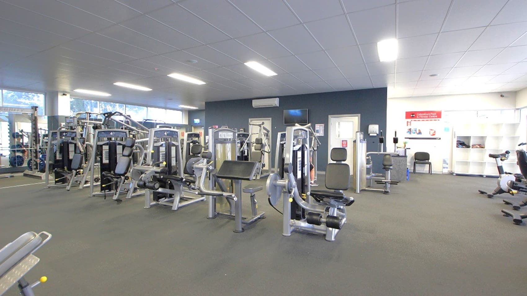 A gym interior with various exercise machines arranged in rows on a grey carpeted floor. On the left, there are weight machines and cardio equipment near large windows. In the centre, more weight machines are visible. On the right, there is a wall with a red sign and a shelving unit with cubbies. The ceiling is fitted with rectangular fluorescent lights, and the walls are painted in neutral tones.