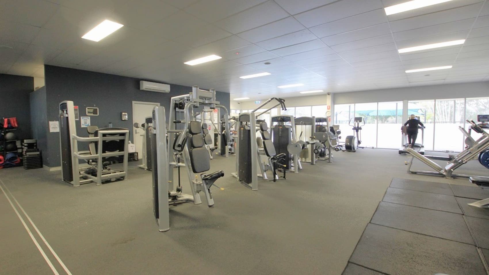 A gym interior with various exercise machines. On the left, there are weight machines and a dark wall with framed pictures. In the centre, more weight machines are arranged in rows. On the right, a person is using a treadmill near large windows that let in natural light. The floor is carpeted, and the ceiling has rectangular fluorescent lights.