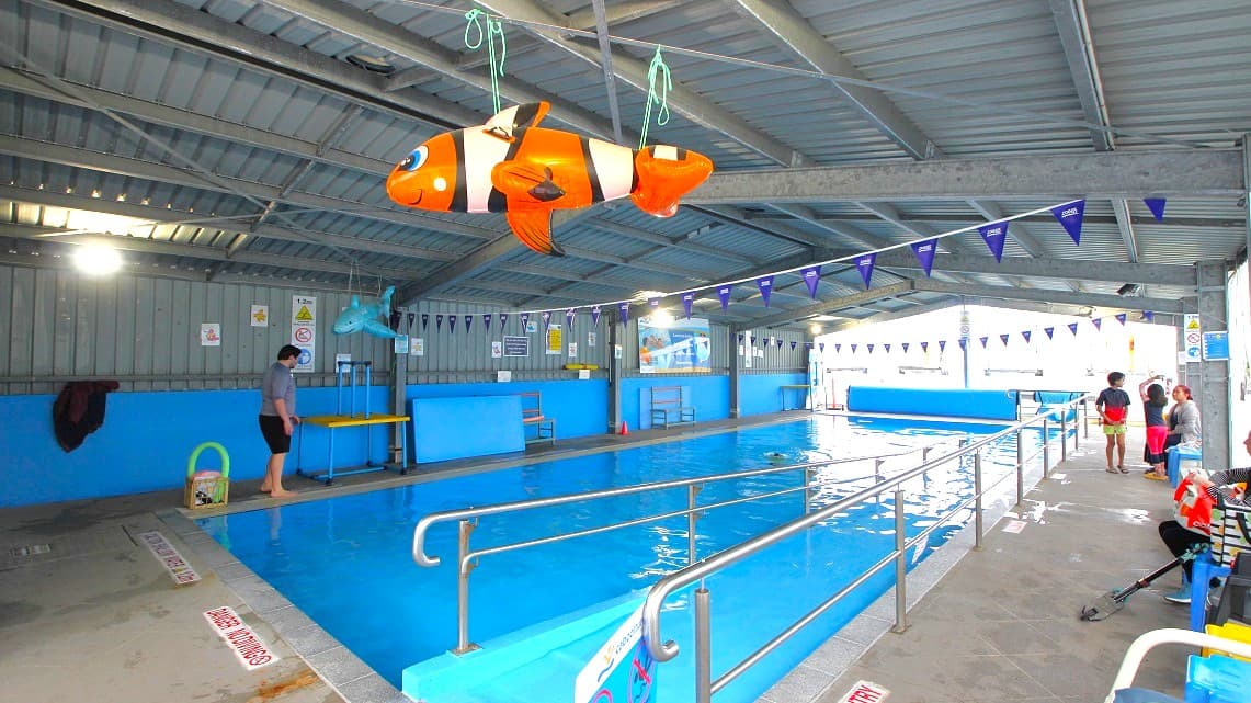 An indoor swimming pool with a metal roof and blue walls. A large inflatable clownfish is suspended from the ceiling. On the left, a person stands near the pool's edge. The pool is bordered by a metal railing, and triangular flags hang above. On the right, a few people, including a child, are near the poolside. The floor is tiled, and there are various signs on the walls. The pool water is calm, reflecting the interior lights.