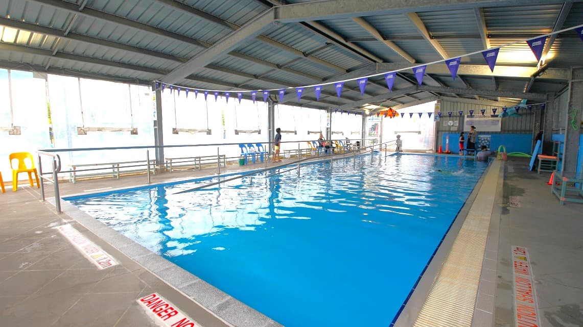 An indoor swimming pool with clear blue water. On the left, there are yellow and blue plastic chairs along the poolside. The ceiling is made of metal beams, and purple triangular flags are strung across the pool. The right side has a tiled walkway with safety warnings. In the background, a few people are near the pool, and there are windows allowing natural light to enter. The floor is concrete with drainage grates along the pool's edge.