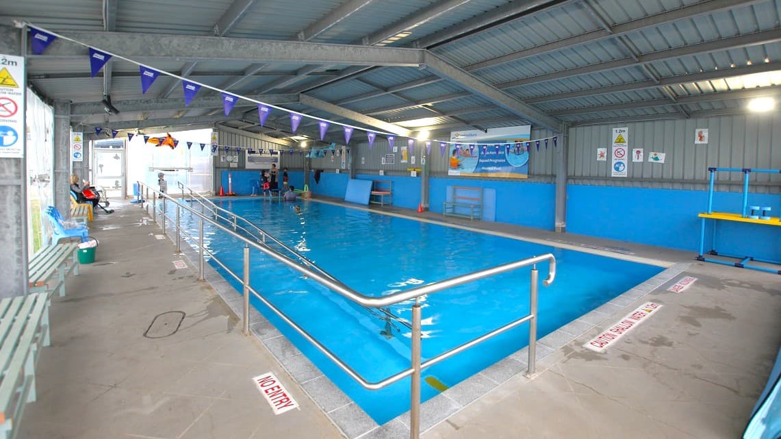 Indoor swimming pool with a metal railing along the edge. The pool is surrounded by blue walls and a corrugated metal ceiling. On the left, there are benches and a few signs, including a "No Entry" sign on the floor. The right side has a shelf with water bottles. Bunting hangs across the pool, and there are a few people in the water. The floor is wet, and the area is well-lit with overhead lights.