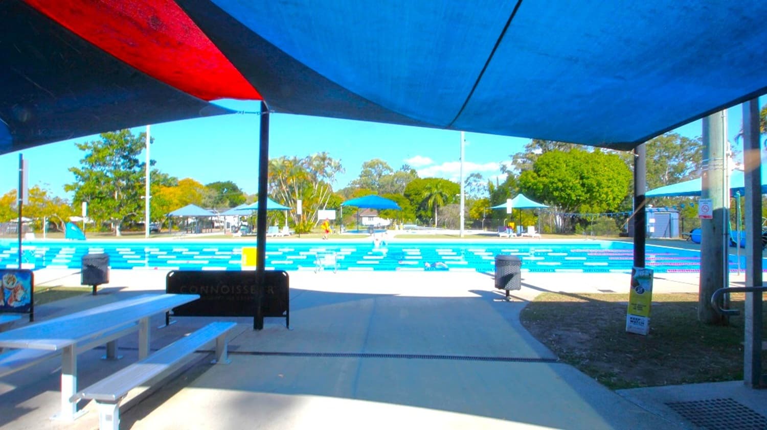 A swimming pool with blue and white lane dividers is in the centre, surrounded by a paved area. Blue shade structures cover parts of the seating area in the foreground, which includes white benches and a black bin. In the background, there are trees and more blue shade structures. The sky is not visible.