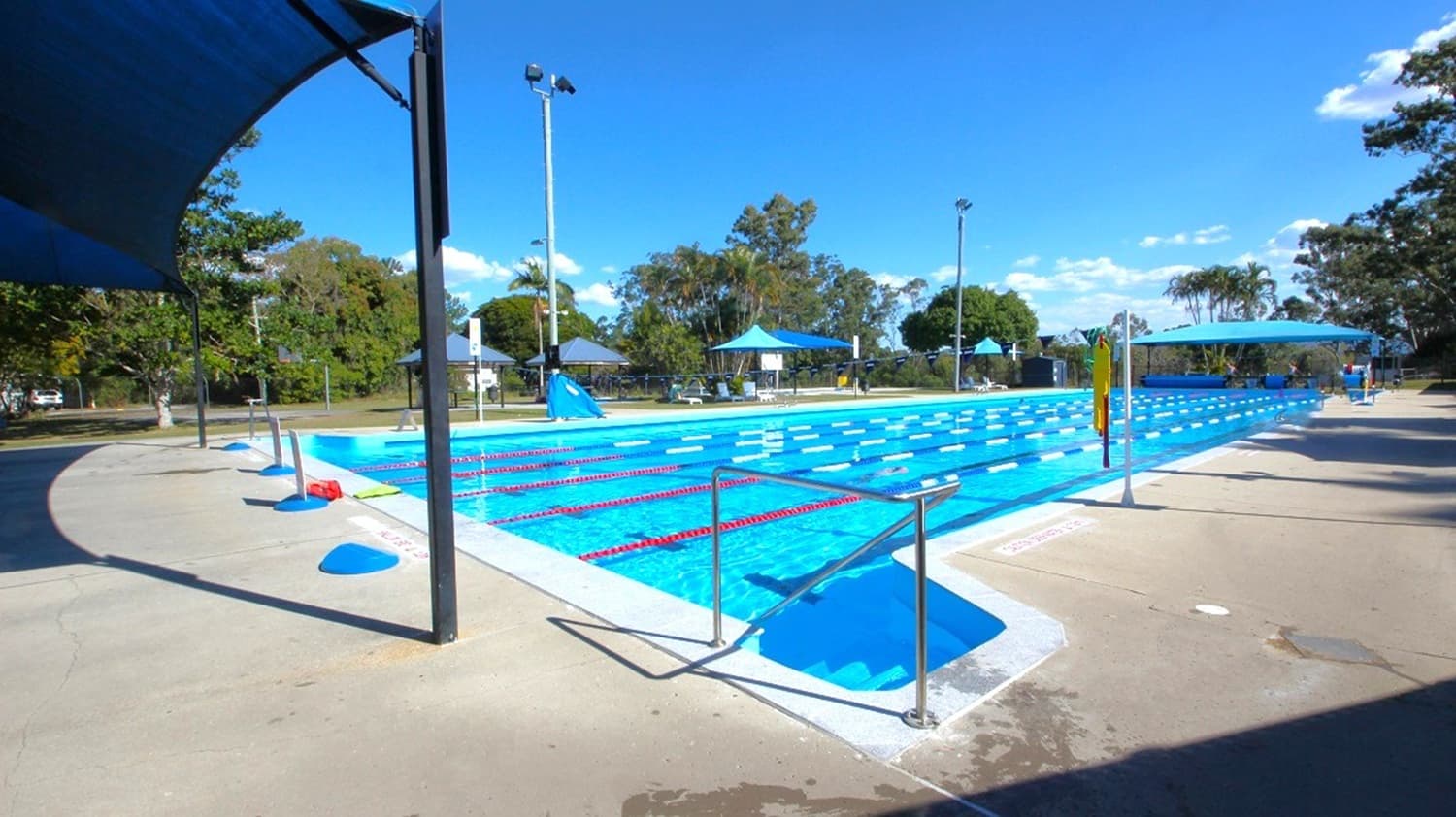 An outdoor swimming pool with clear blue water and lane dividers. On the left, a shaded area with a canopy and a concrete walkway. In the background, trees and a few small structures with blue roofs. On the right, a person is walking along the poolside. The pool is surrounded by a concrete deck, and there are tall lights around the area. The sky is clear and blue.