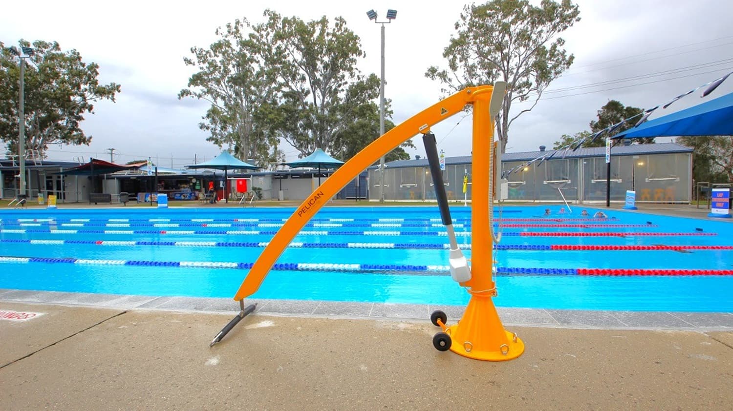 A swimming pool with multiple lanes marked by red and blue lane dividers. In the foreground, a yellow pool lift is positioned on the pool deck, designed to assist individuals with mobility challenges. The background features trees and a building with a blue roof. The pool area is surrounded by a fence, and there are some poolside chairs and equipment visible. The sky is overcast.
