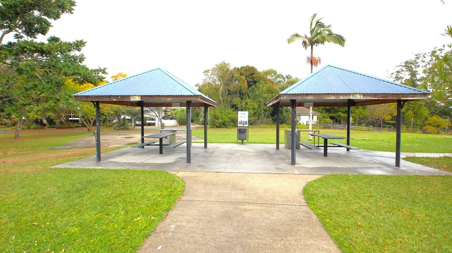 Two picnic shelters with blue metal roofs are in the centre, each covering a picnic table. A concrete path leads to the shelters, surrounded by green grass. In the background, there are trees, including a tall palm tree, and a few parked cars partially visible. A bin and a sign are positioned between the shelters.