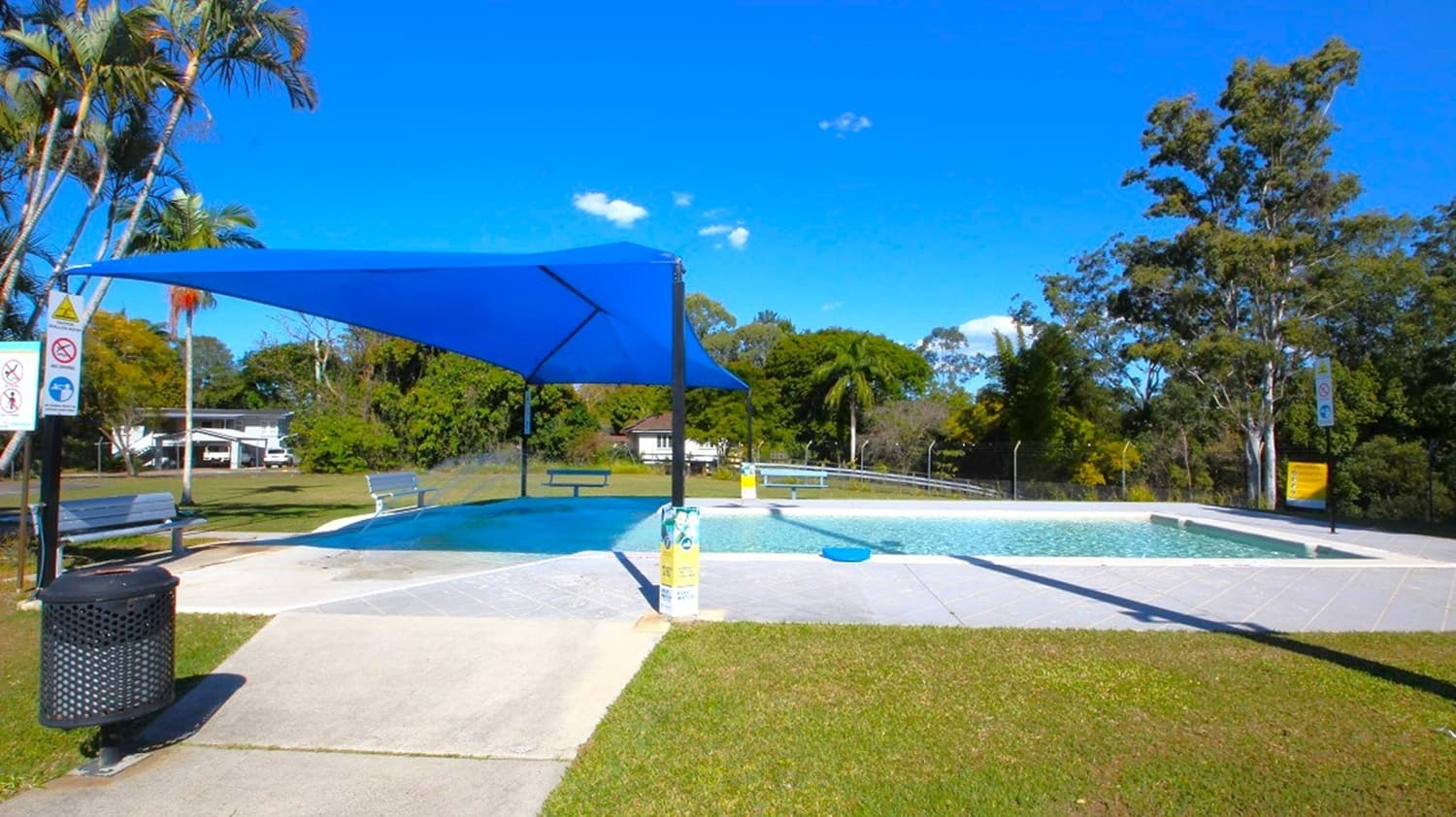 A small outdoor pool with a blue shade sail overhead. The pool is surrounded by a concrete path and grassy areas. On the left, there is a black rubbish bin and a signpost. Benches are placed around the pool area. In the background, there are trees and a few buildings, including a white structure partially visible. The sky is clear with a few clouds.