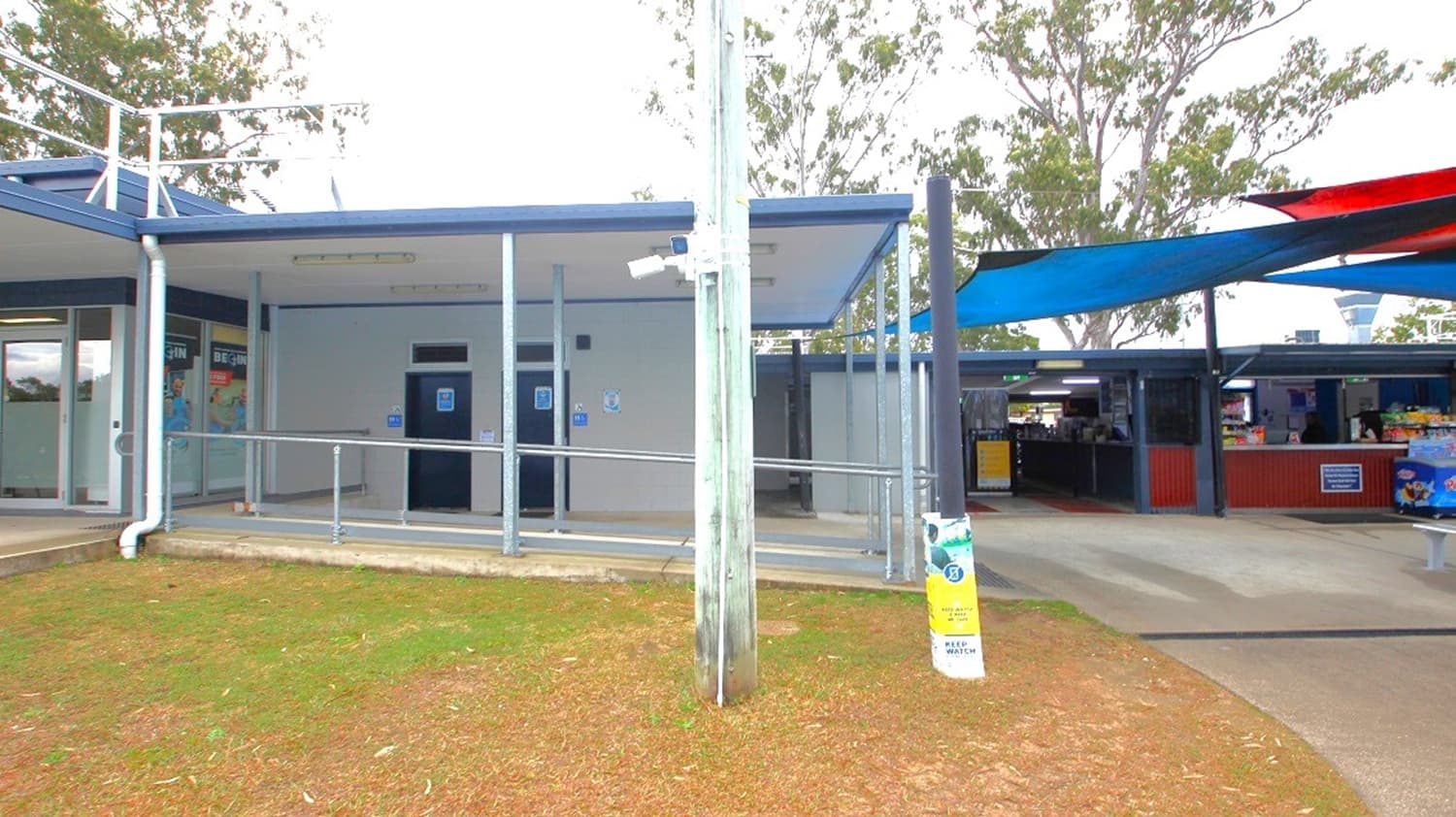 On the left, there is a building with a blue roof and a ramp leading to a door, possibly a public restroom. A white pipe runs down the side. In the centre, a tall pole with a security camera is visible. On the right, there are shaded areas with blue and red canopies, covering an open space with tables and a kiosk. The ground is a mix of grass and concrete, and trees are visible in the background.