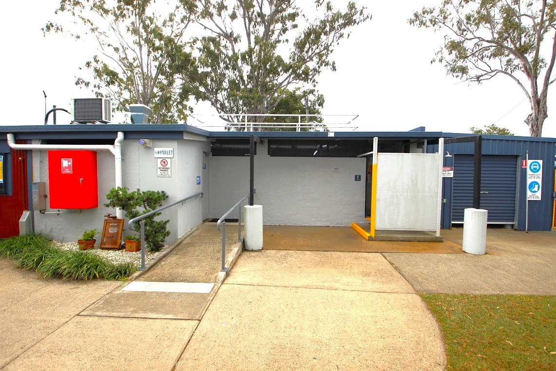 A public restroom facility with a grey exterior. On the left, there's a red fire hose reel mounted on the wall, with a small garden area featuring plants and a sign. A ramp with a metal railing leads to the entrance. The middle section has a grey wall with a door, and a yellow bollard near the entrance. On the right, there are additional signs and a dark blue section of the building. Trees are visible in the background, and the ground is paved with concrete.