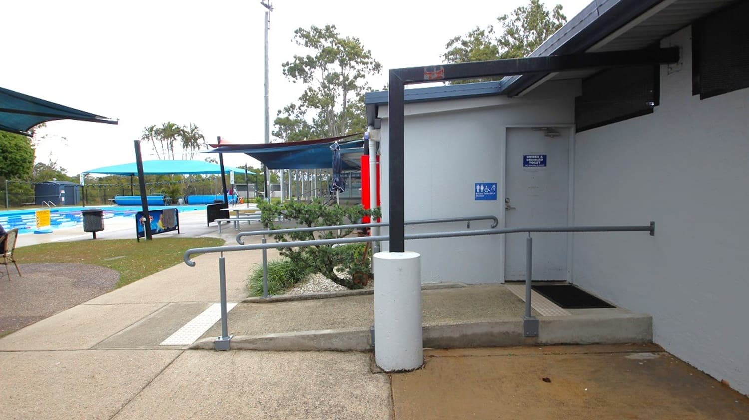 A swimming pool area with a pathway leading to a building on the right. The building has a ramp with handrails and a door marked with a sign. On the left, there are shaded seating areas with blue canopies, tables, and chairs. The pool is visible in the background, with lanes and a few people swimming. Trees and a fence are in the distance. The ground is a mix of concrete and grass.