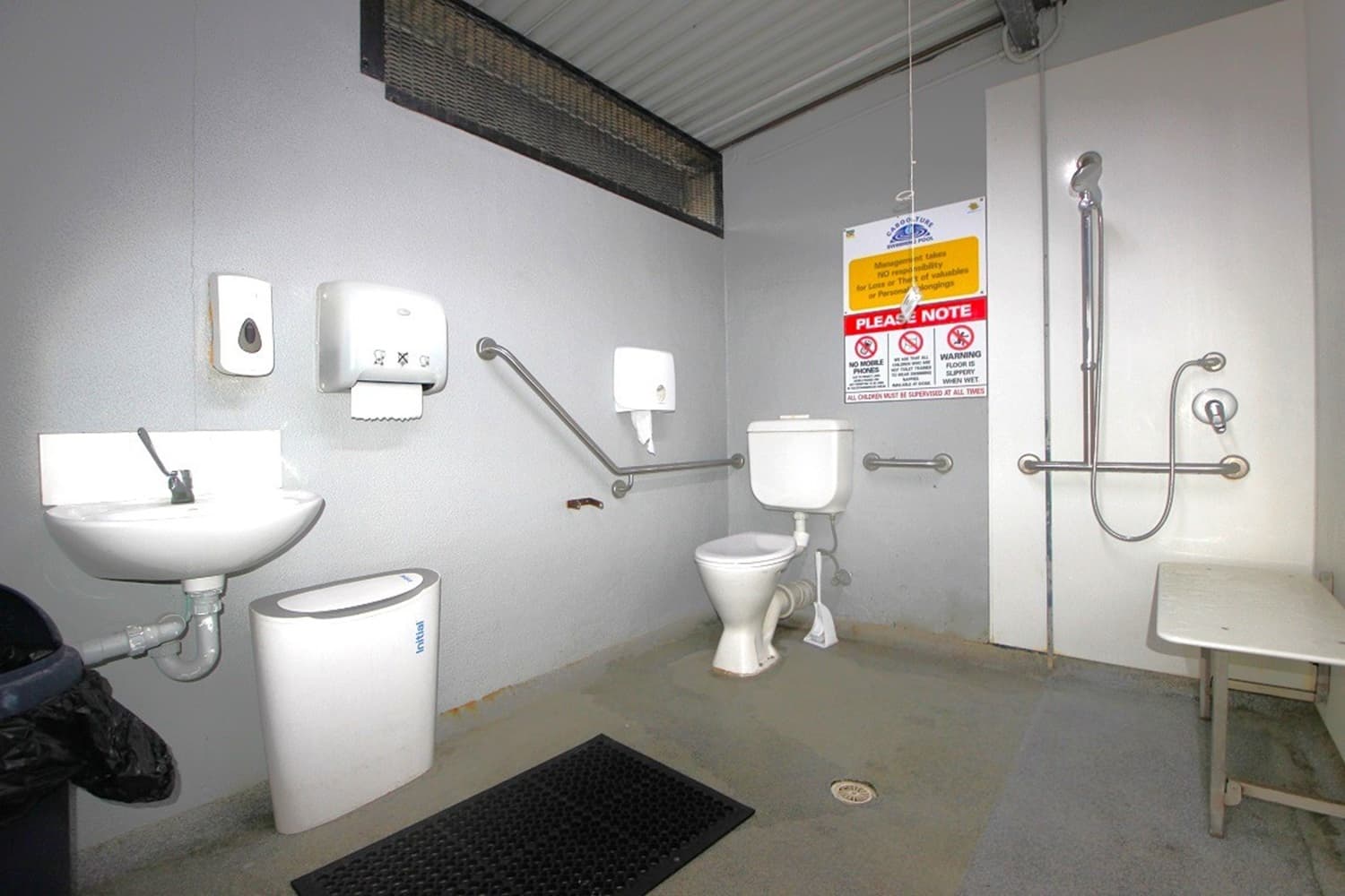 A public restroom with accessibility features. On the left, a small sink with a soap dispenser and hand dryer above it. A sanitary bin is below. In the centre, a toilet with grab bars on both sides. Above the toilet, a sign with usage instructions. On the right, a shower area with a handheld showerhead and additional grab bars. A foldable shower seat is attached to the wall. The floor is non-slip, and the ceiling is corrugated metal.