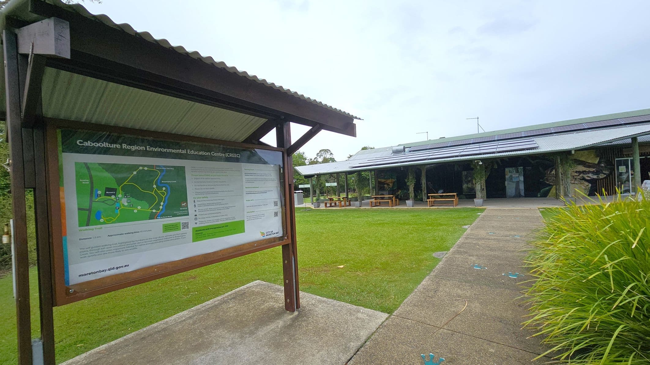 A covered information board on the left displays a map and details about the Caboolture Region Environmental Education Centre. The board is supported by wooden posts. On the right, a building with a corrugated metal roof and solar panels is visible, featuring open sides and picnic tables. The foreground has a paved path leading to the building, bordered by green grass and plants.