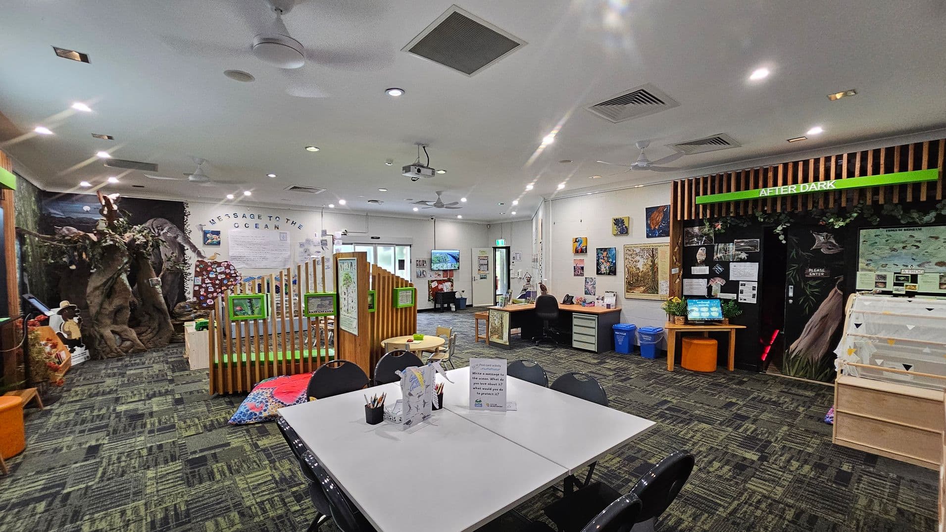 A spacious room with a green and grey patterned carpet. On the left, there's a large tree structure and a wooden fence with green signs. In the centre, a table with chairs and art supplies. The back wall has a "Message to the Ocean" display. On the right, a section labelled "After Dark" with a computer and informational posters. The ceiling is white with recessed lighting and ceiling fans. Walls are adorned with various artworks and educational materials.