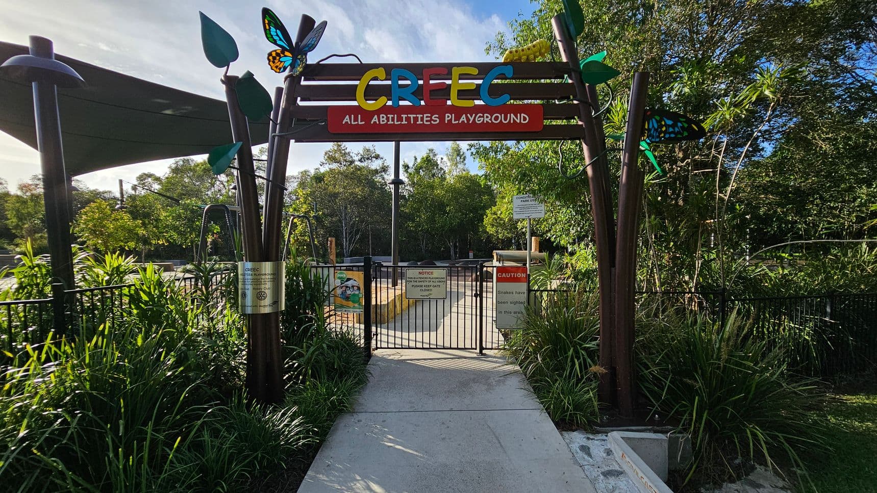 Entrance to a playground with a colourful sign reading "CREEC All Abilities Playground" at the top. The gate is black metal with various signs, including a caution about snakes. Decorative elements like butterflies and leaves adorn the entrance. Lush greenery surrounds the area, with trees and plants on both sides. A shaded area with a canopy is visible on the left. The path leading into the playground is concrete.