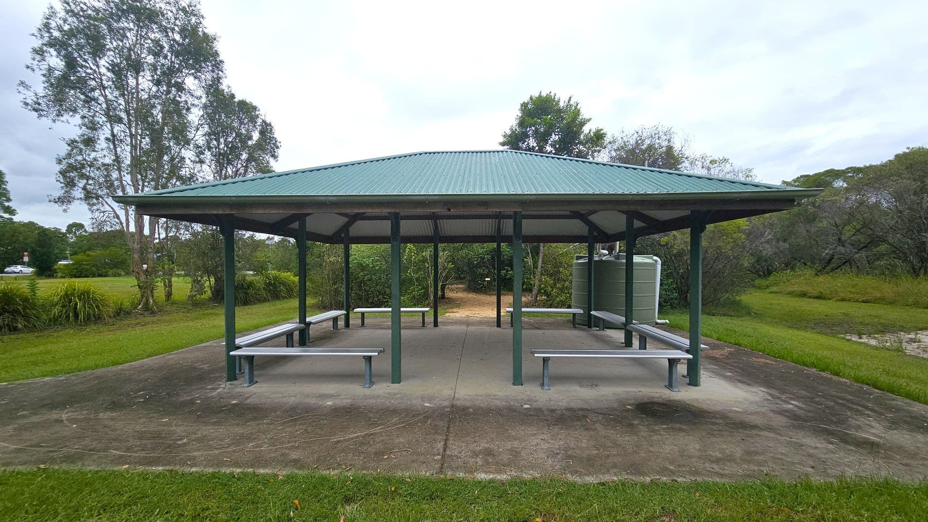 A green-roofed pavilion with open sides and several benches underneath. The structure is supported by green metal poles and stands on a concrete slab. In the background, there are trees and bushes, with a grassy area surrounding the pavilion. A water tank is visible on the right side. The ground is a mix of grass and concrete, and the area appears to be part of a park or recreational space.