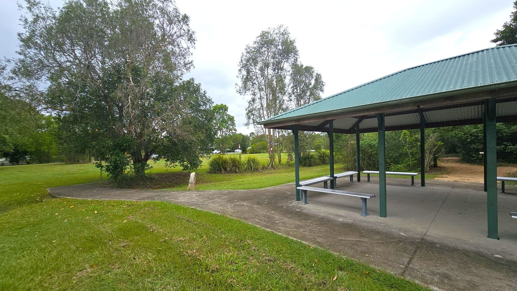 A park scene with a green-roofed shelter on the right, featuring benches underneath. The ground is paved beneath the shelter, transitioning to grass. On the left, there are large trees with dense foliage and a grassy area. In the background, more trees and open grassy fields are visible, with a few shrubs near the shelter. The sky is overcast.