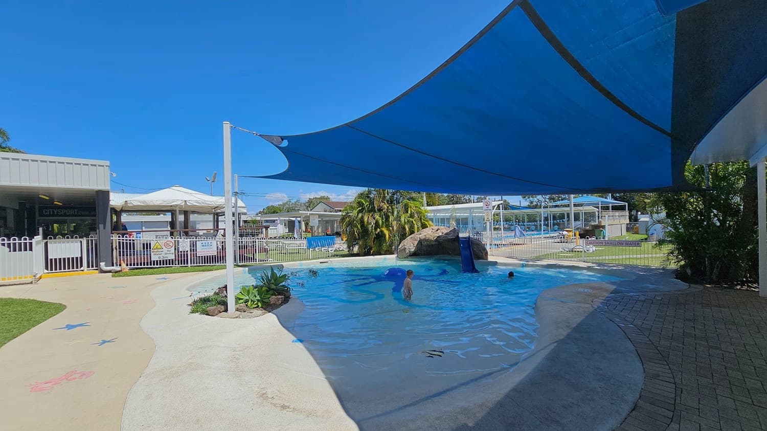 A shallow outdoor pool with a blue shade sail overhead. On the left, there is a fenced area with a sign that reads "City Sport" and a white gate. The pool has a small slide attached to a rock structure in the middle. There are a few children playing in the water. The ground around the pool is a mix of concrete and brick paving. In the background, there are more fenced areas, a grassy lawn, and some buildings with white roofs.