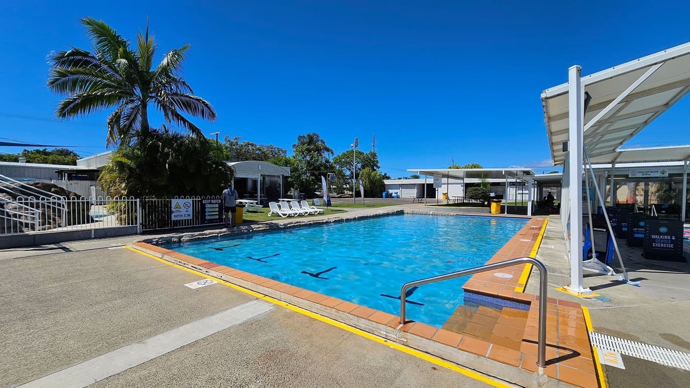 Outdoor swimming pool with clear blue water. On the left, there is a white fence, a palm tree, and several white sun loungers on a grassy area. In the middle, the pool is bordered by a brick edge and a metal handrail leading into the water. On the right, there is a covered area with a white roof and signs indicating "Walking & Gentle Exercise." The ground around the pool is a mix of concrete and textured surfaces.