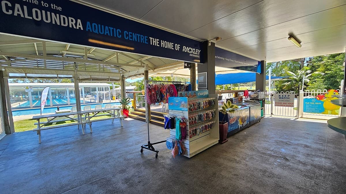 A covered area with a sign reading "Caloundra Aquatic Centre" at the top. On the left, there are picnic tables and a view of a swimming pool with lanes. In the middle, there is a rack displaying swimwear and accessories. On the right, there is a counter with a freezer and a sign that says "Keep Watch Around Pools." The floor is concrete, and the ceiling is white with fluorescent lighting. In the background, there are trees and a fence with a gate.