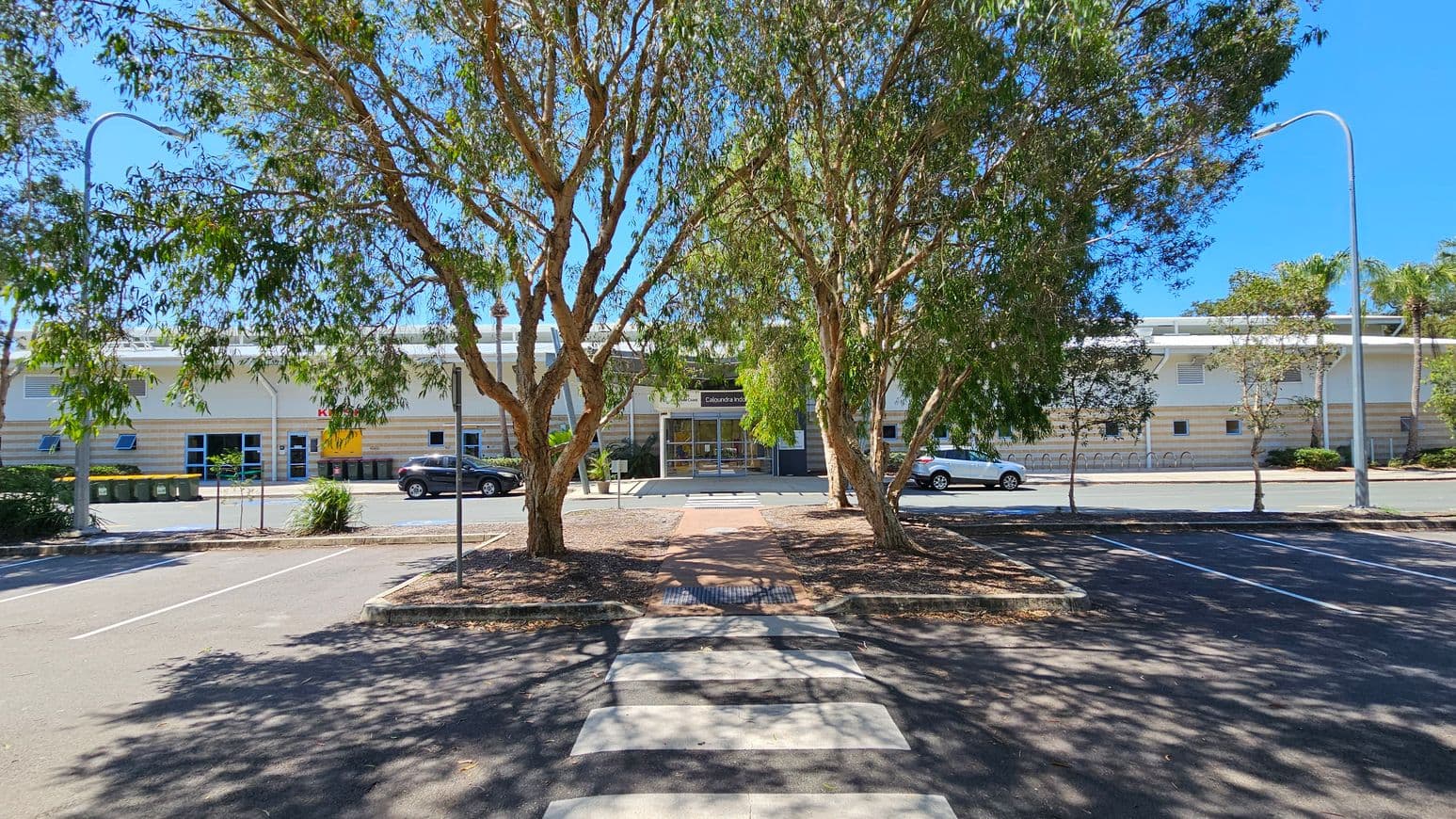 A car park with a zebra crossing in the foreground leads to a building in the background. The building has a modern design with a glass entrance and a sign above the door. Two large trees with green foliage are in the centre, casting shadows on the ground. Cars are parked along the street in front of the building. Street lamps are visible on both sides, and there are several bins to the left. The sky is clear and blue.